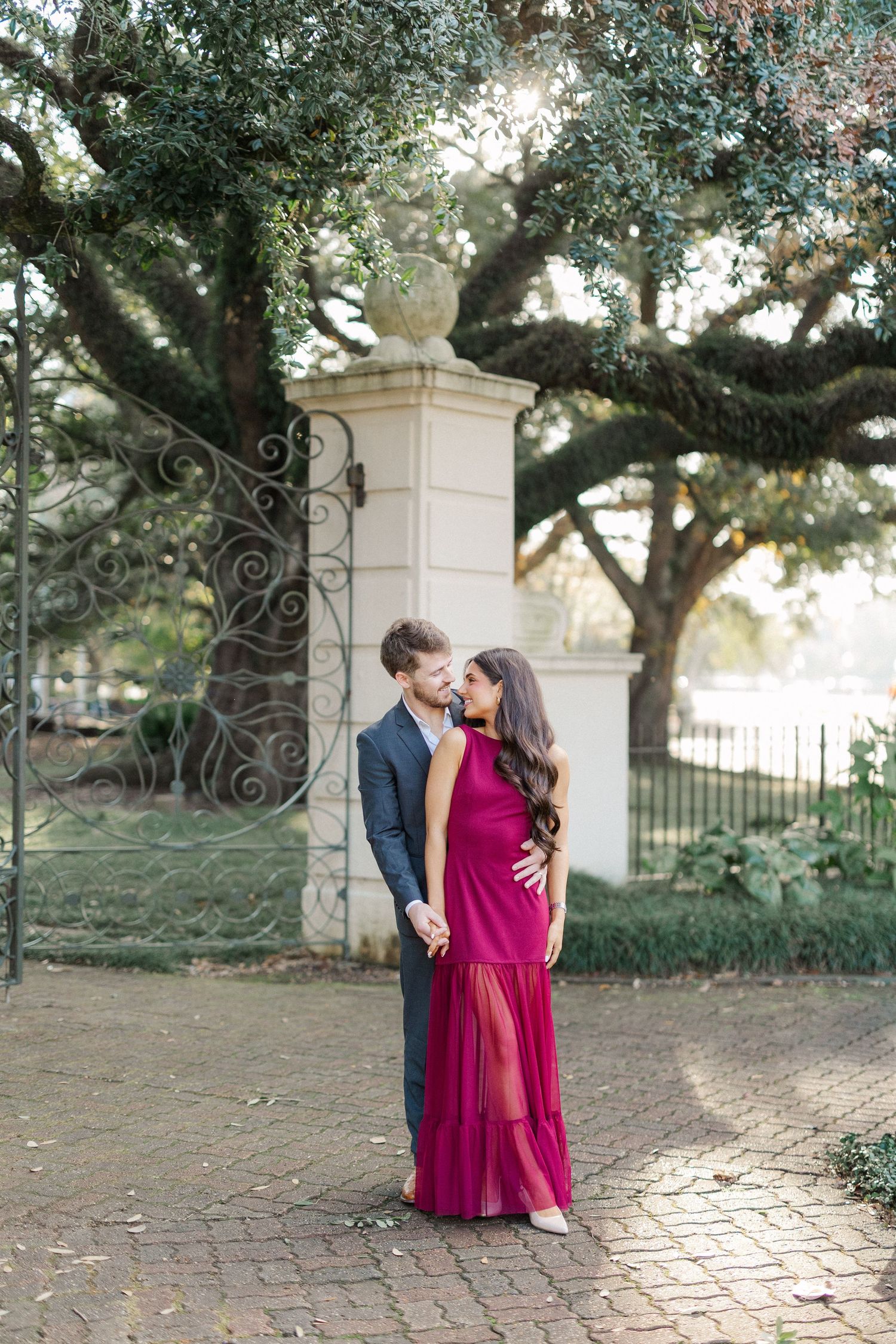 A romantic couple poses in front of an ornate garden gate with Spanish moss draped trees overhead.