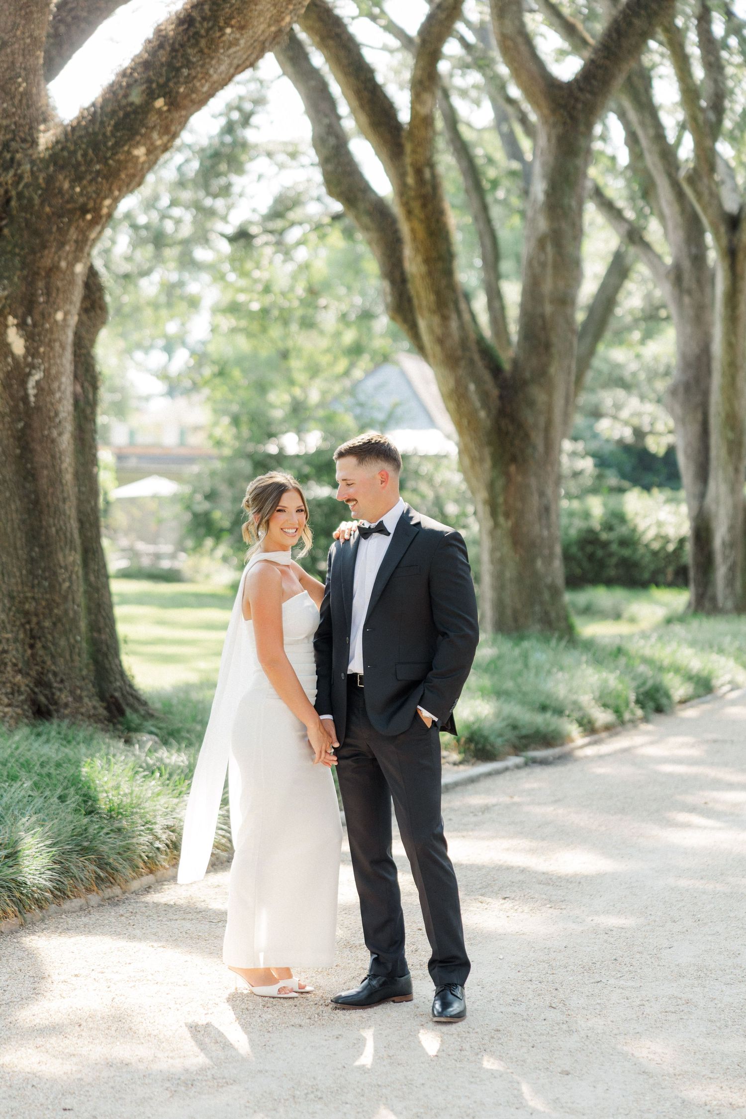 A couple in wedding attire shares a romantic moment under a canopy of oak trees on a sunlit garden path.