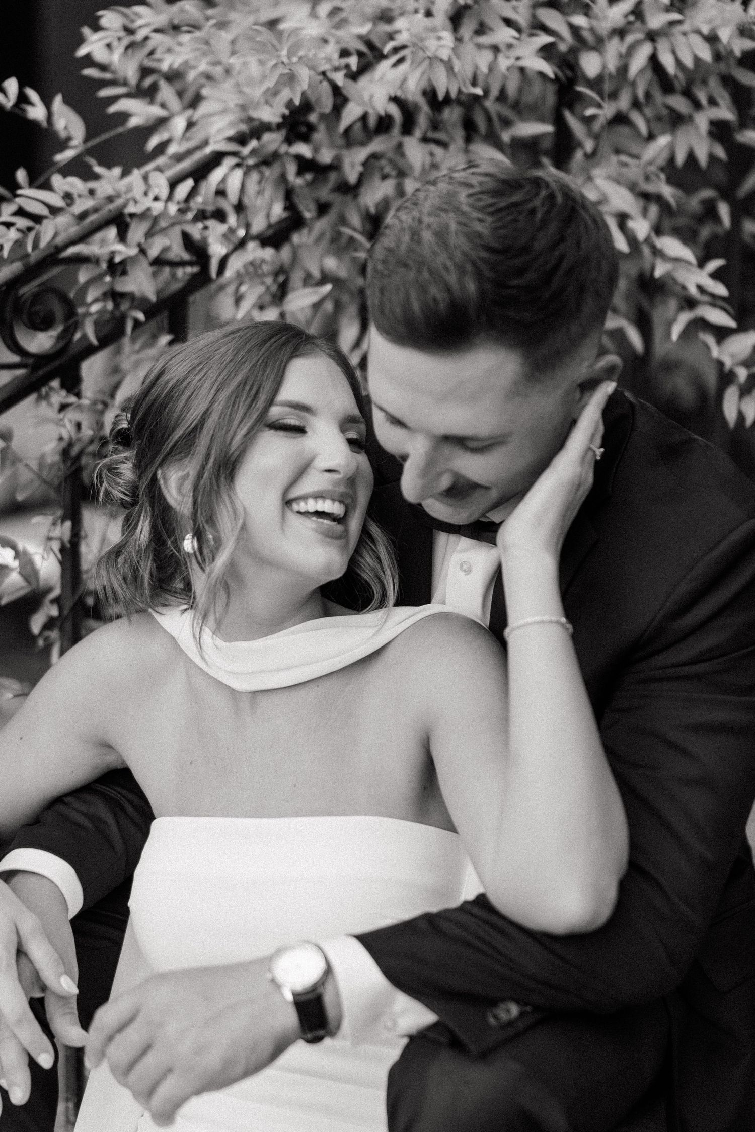 A joyful black and white wedding photo capturing a candid moment between newlyweds against foliage.
