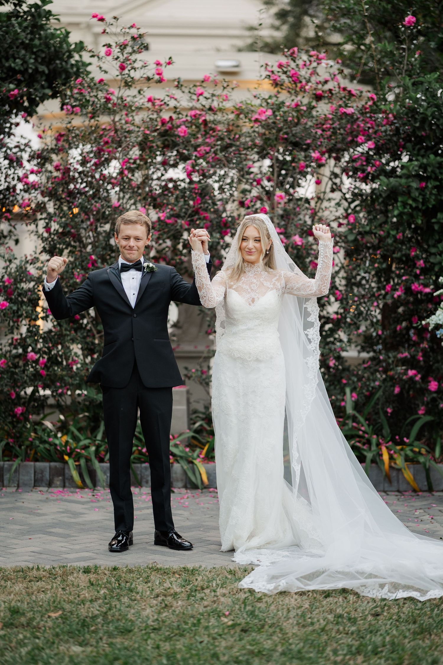 A celebratory wedding pose in front of blooming pink roses and garden landscaping.