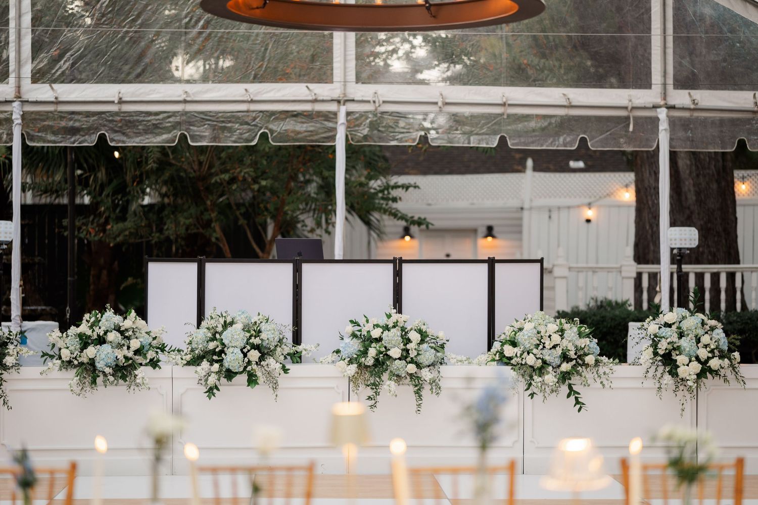 Wedding reception setup with white floral arrangements and modern decor under clear tent ceiling.