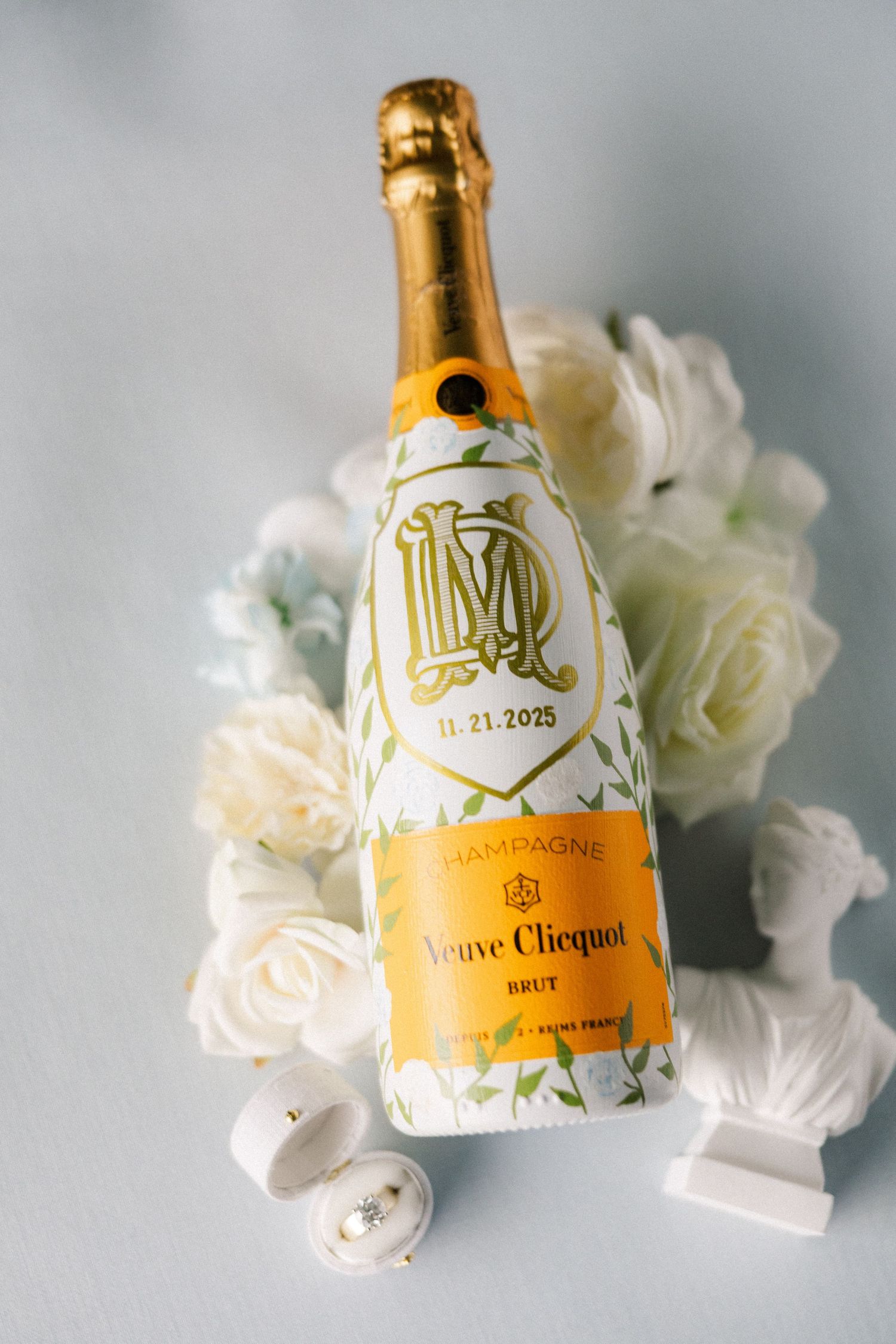 A bottle of Veuve Clicquot champagne with white roses and wedding rings on a light background.
