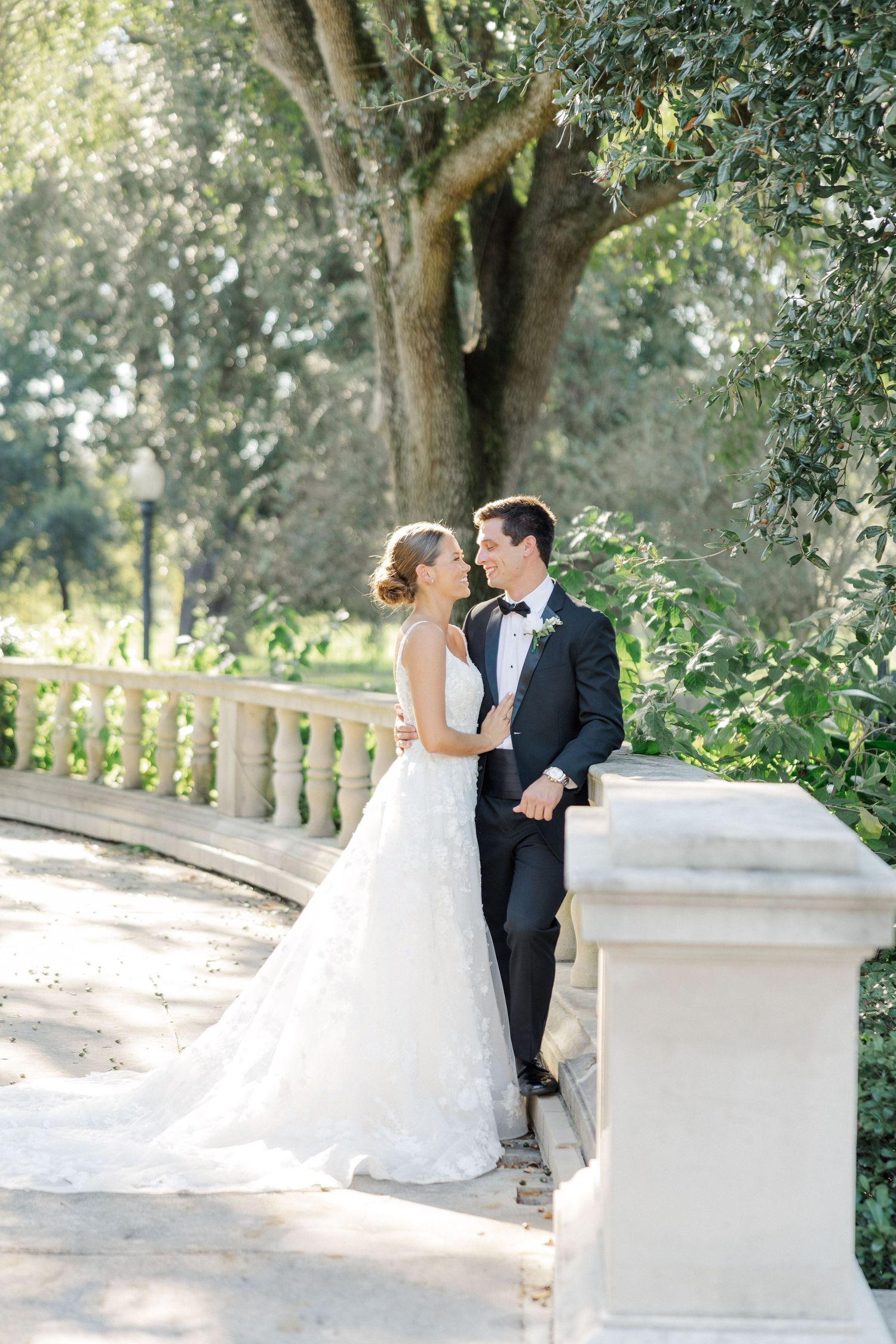 Elegant wedding couple posing along a white stone balustrade in a sunlit garden setting.
