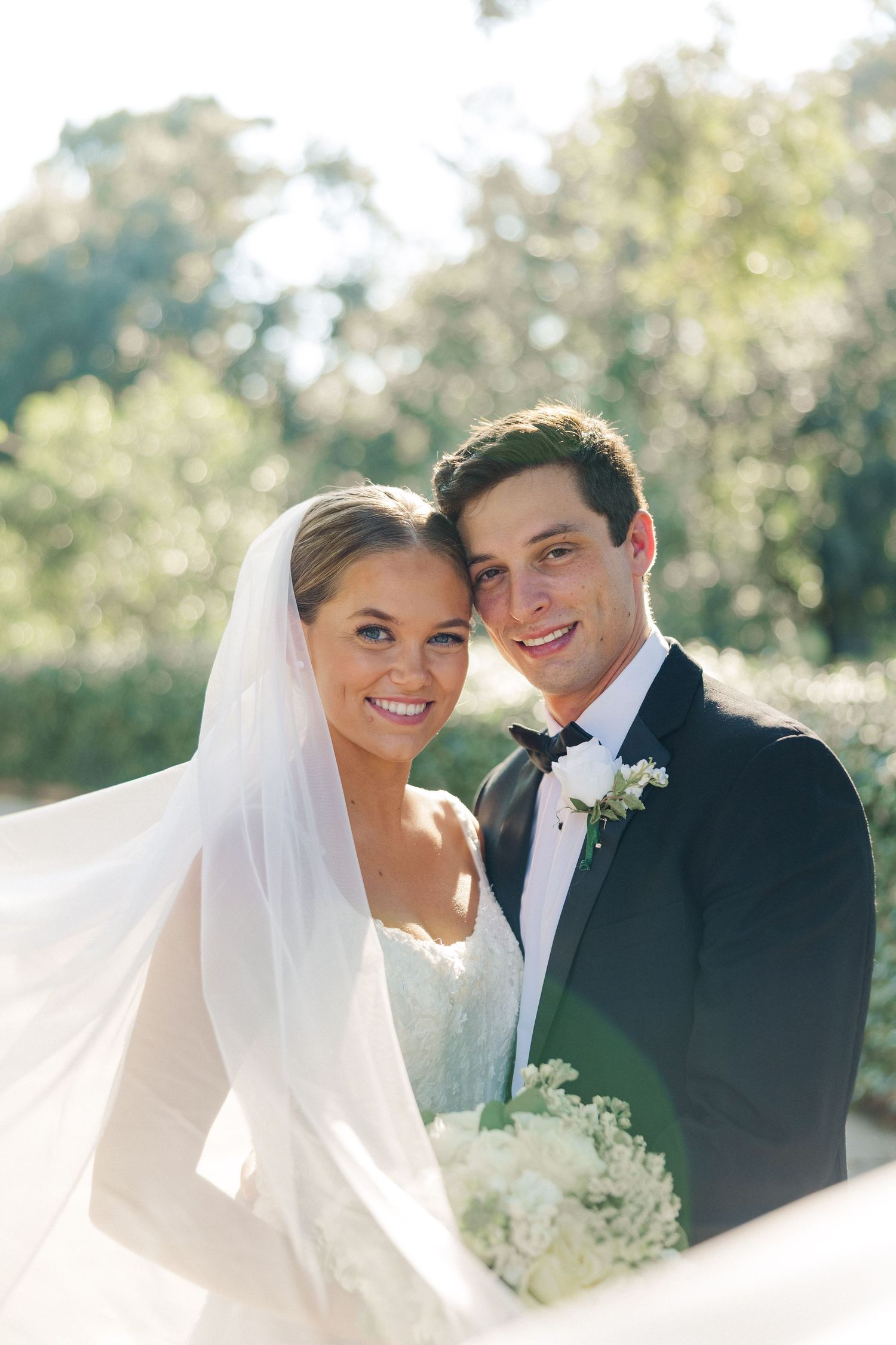 Romantic wedding portrait with bride in white veil and groom in black tuxedo against natural backdrop.