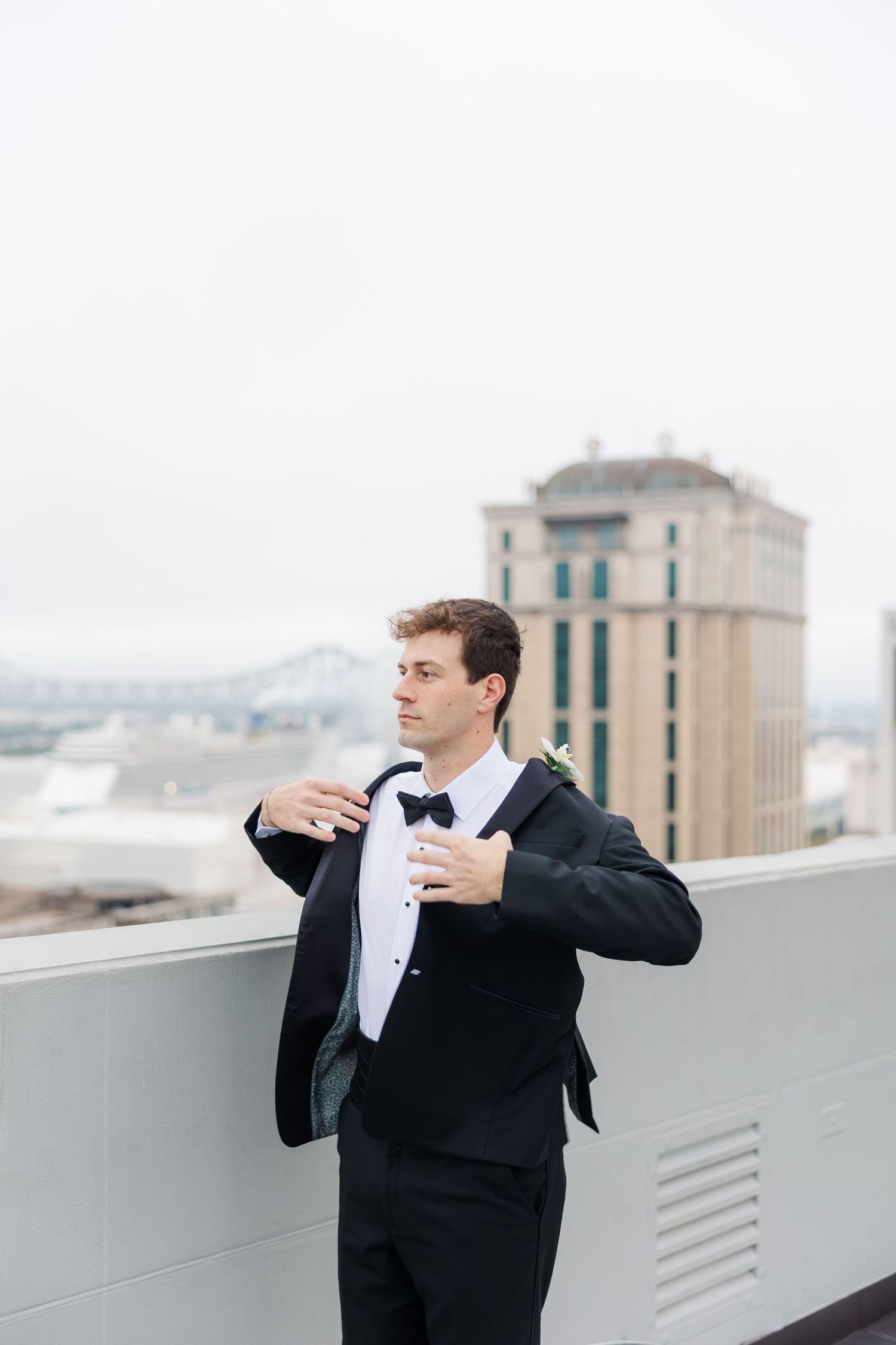 Groom in black tuxedo adjusting his bowtie on a rooftop terrace.