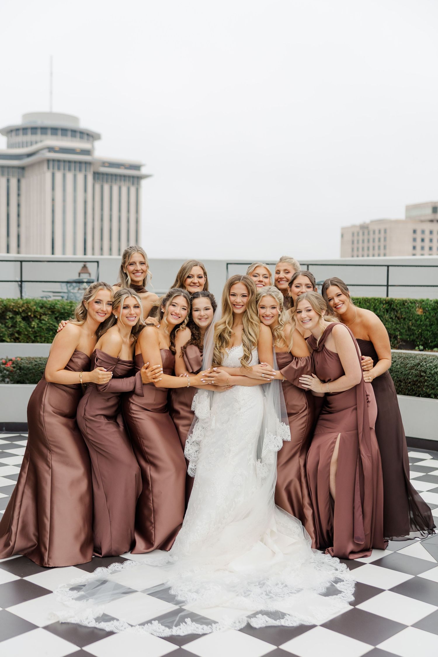 Wedding party in brown dresses posing together on a rooftop with checkerboard floor.