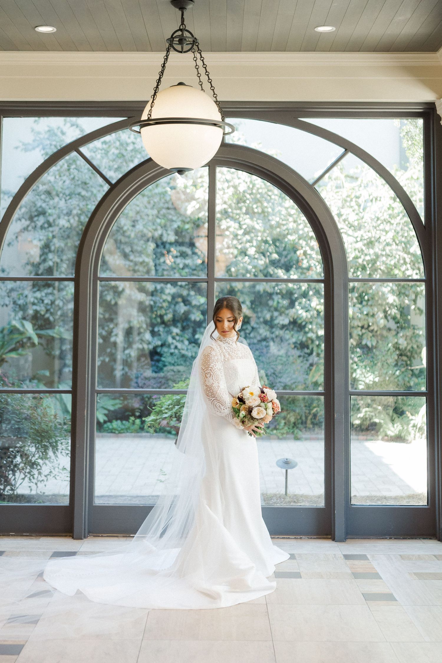 A bride in a long white lace gown and veil stands in front of an elegant arched window with natural light streaming in.
