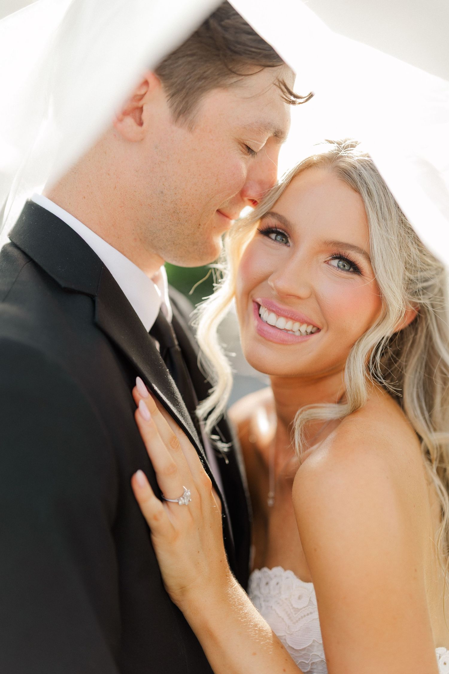 Bride and groom share a romantic moment under a veil, bride smiling with diamond ring visible.