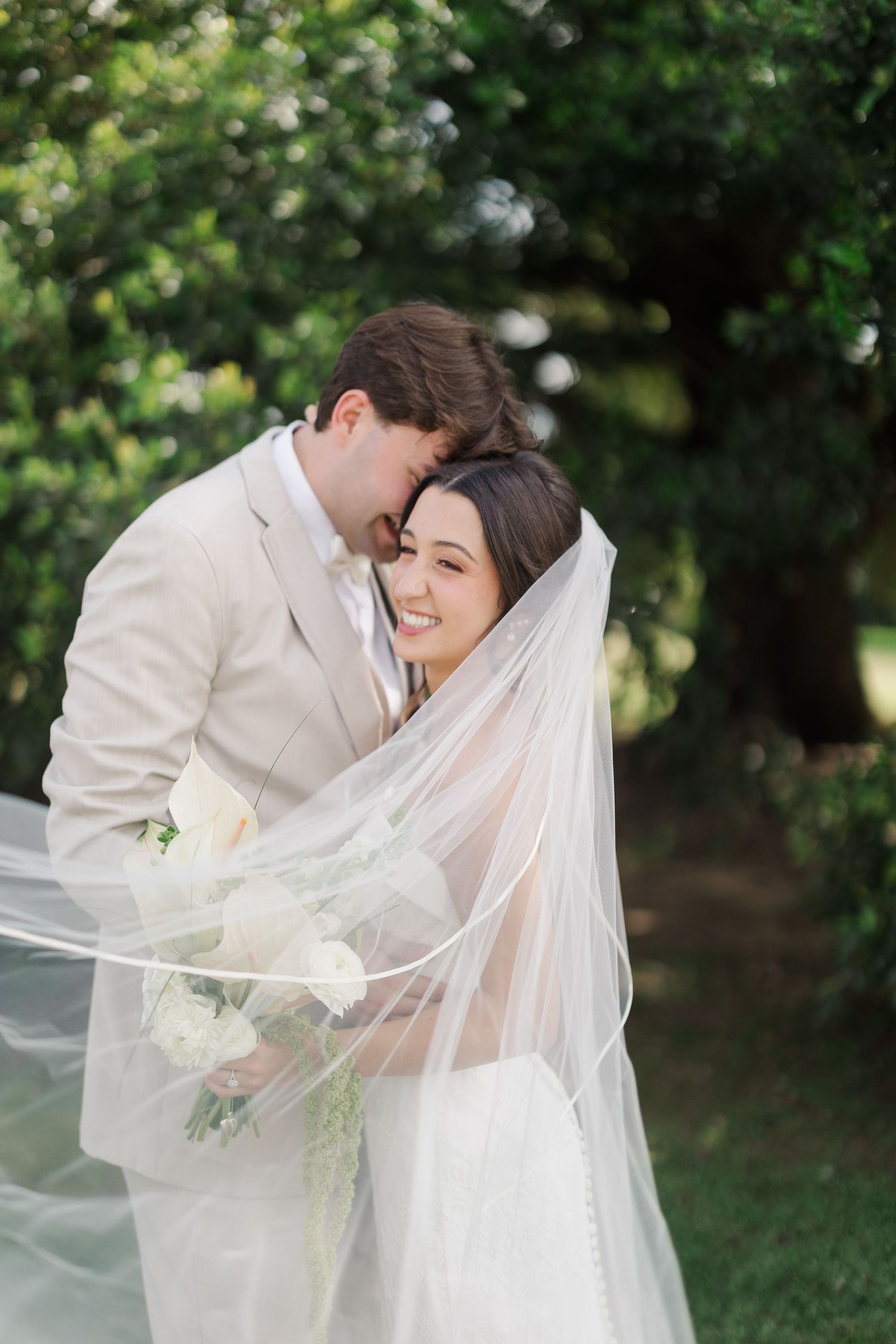 Bride smiling under flowing veil held by groom in tan suit outdoors with lush green trees.
