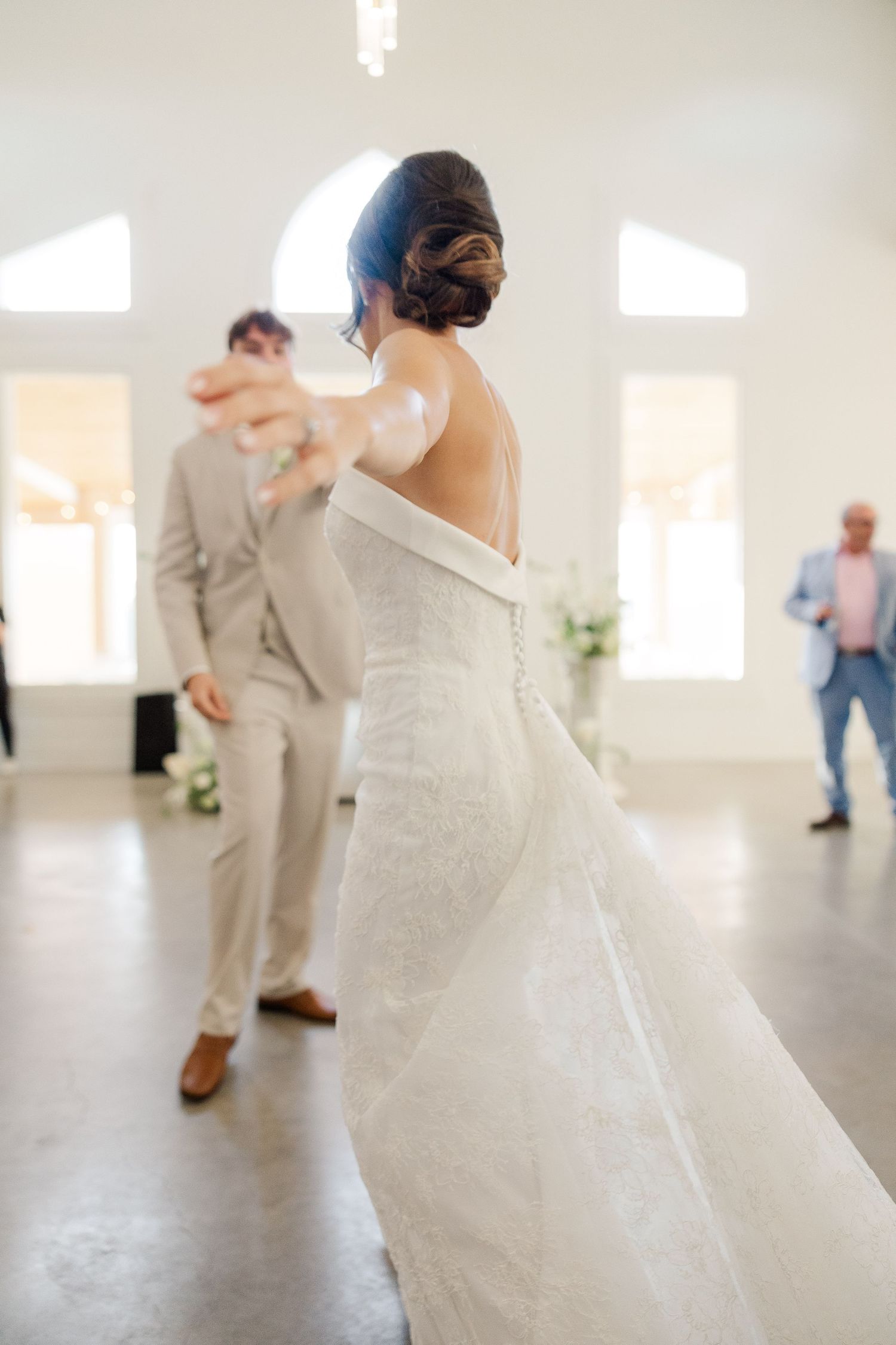 Bride in backless lace gown reaches toward groom during first dance in bright white wedding venue.
