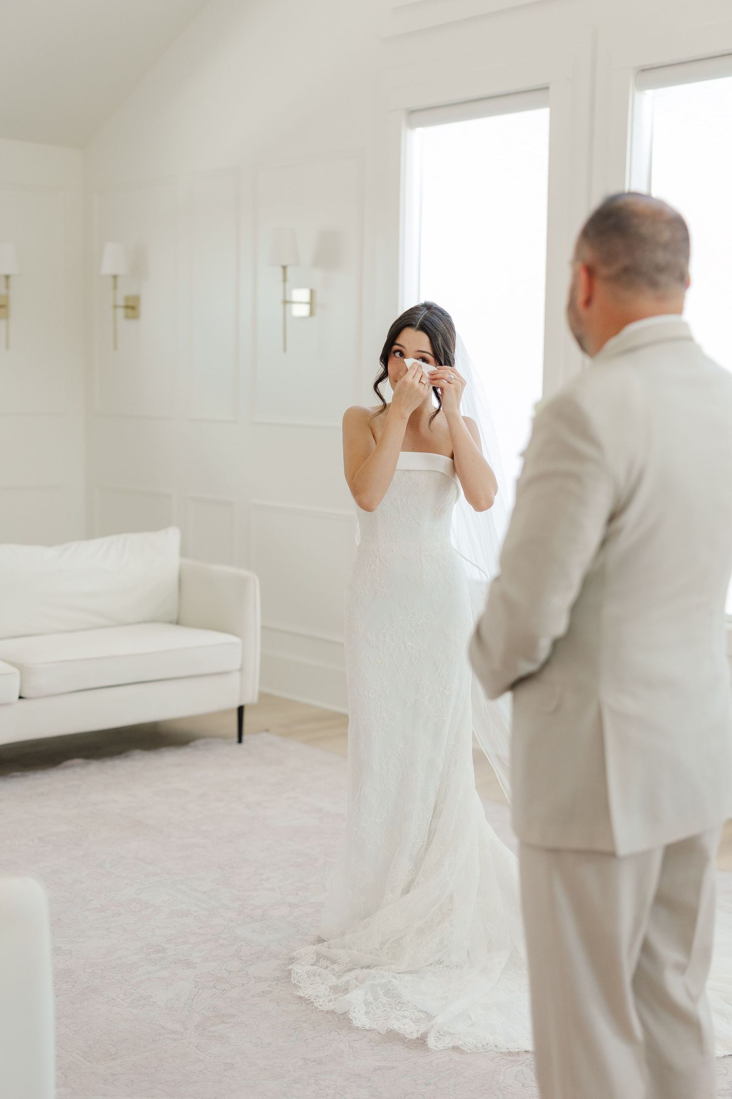 Emotional bride in strapless white gown crying during first look with father in elegant white room.