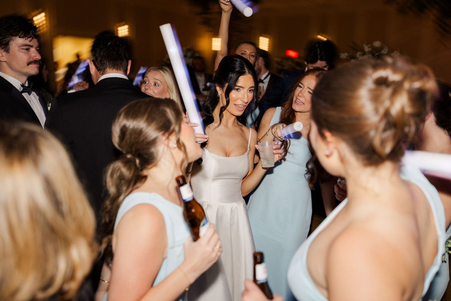 Bride in white gown celebrates on dance floor with bridesmaids holding glow sticks at reception.