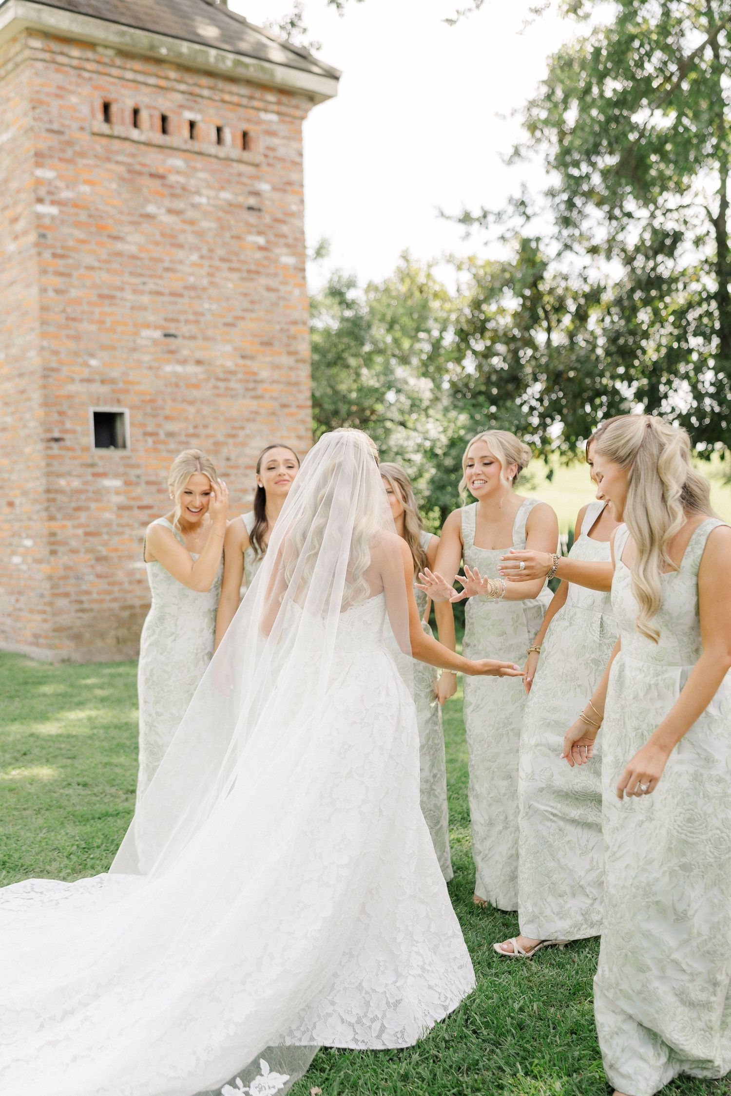 Bride in lace wedding gown with veil surrounded by bridesmaids in white lace dresses near brick tower.