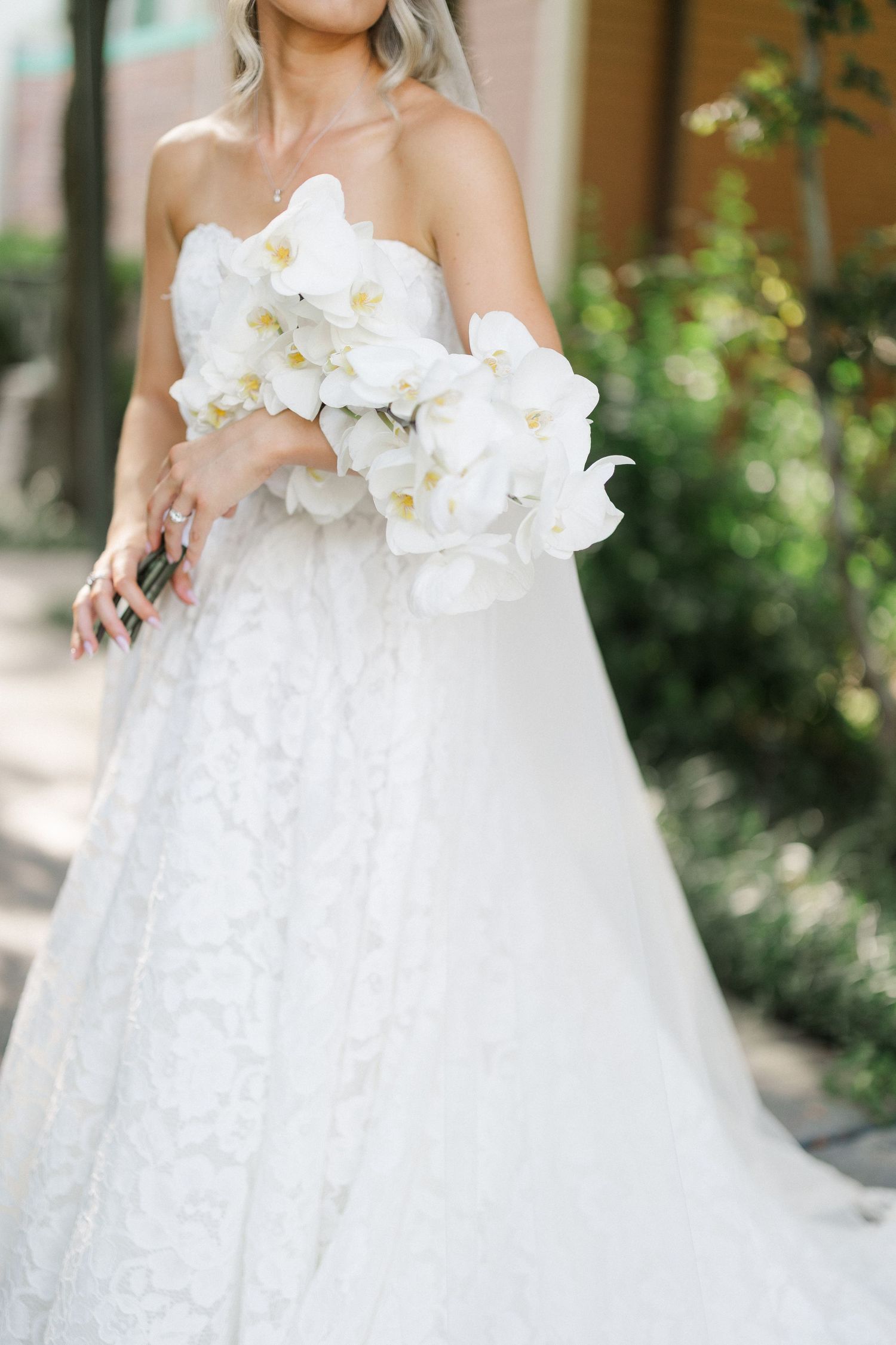 Bride in strapless lace wedding gown holds a stunning white orchid bouquet in an outdoor garden setting.