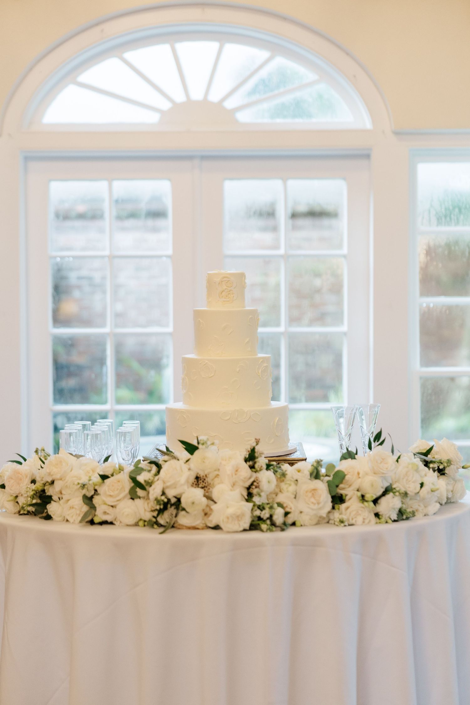 White wedding cake display with floral decorations on table against arched windows in elegant venue.