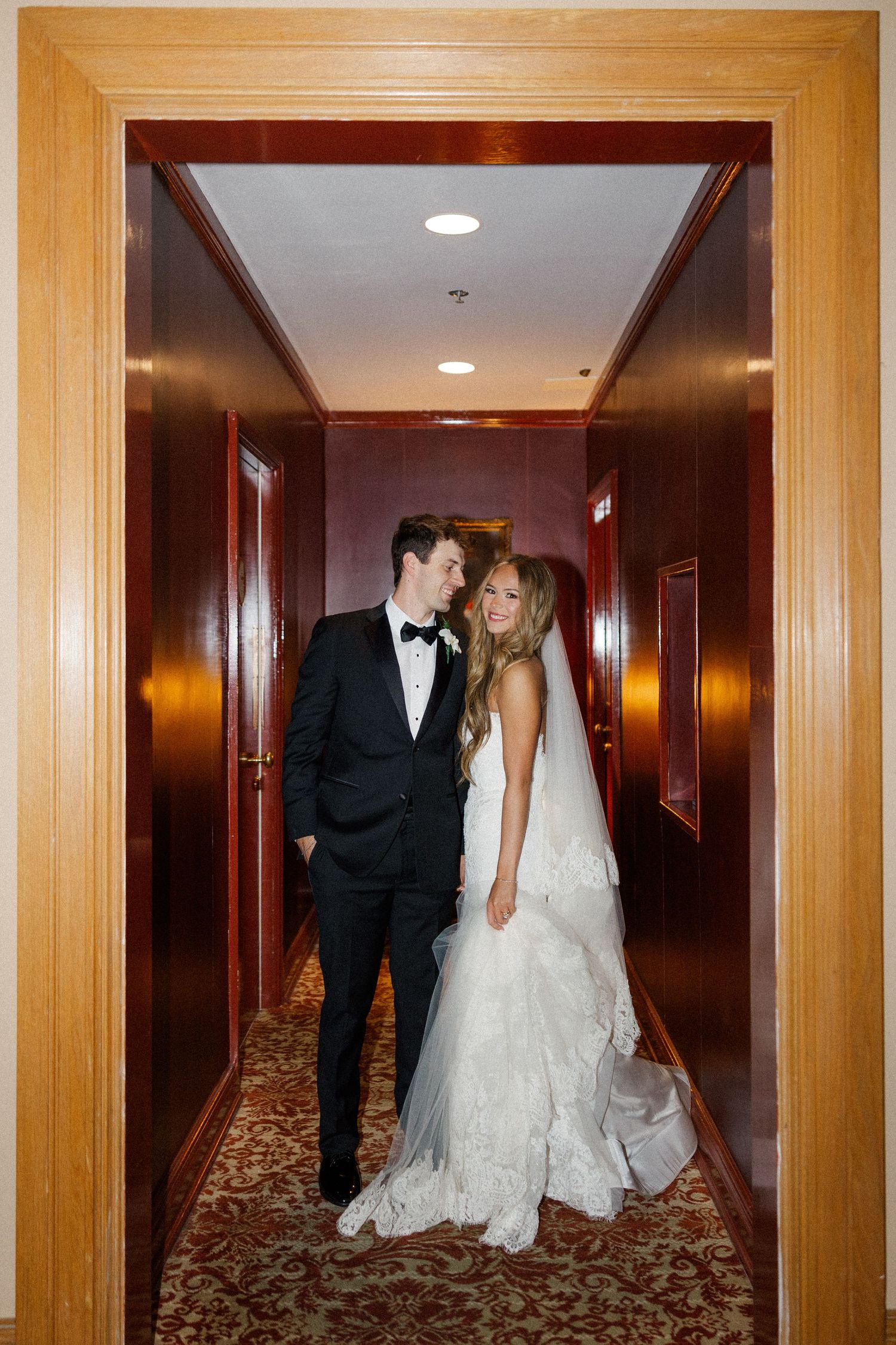 A couple poses in a luxurious hotel corridor with warm wooden walls and ornate carpeting.