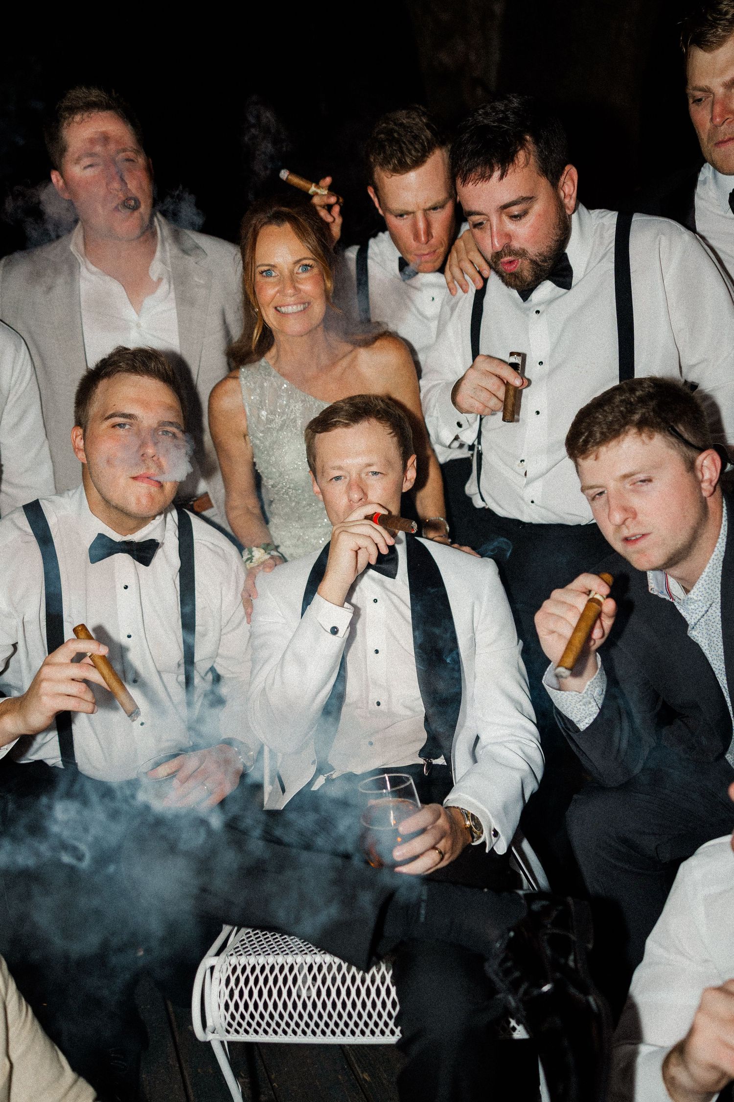 Group of wedding guests in formal attire smoking cigars at evening celebration.