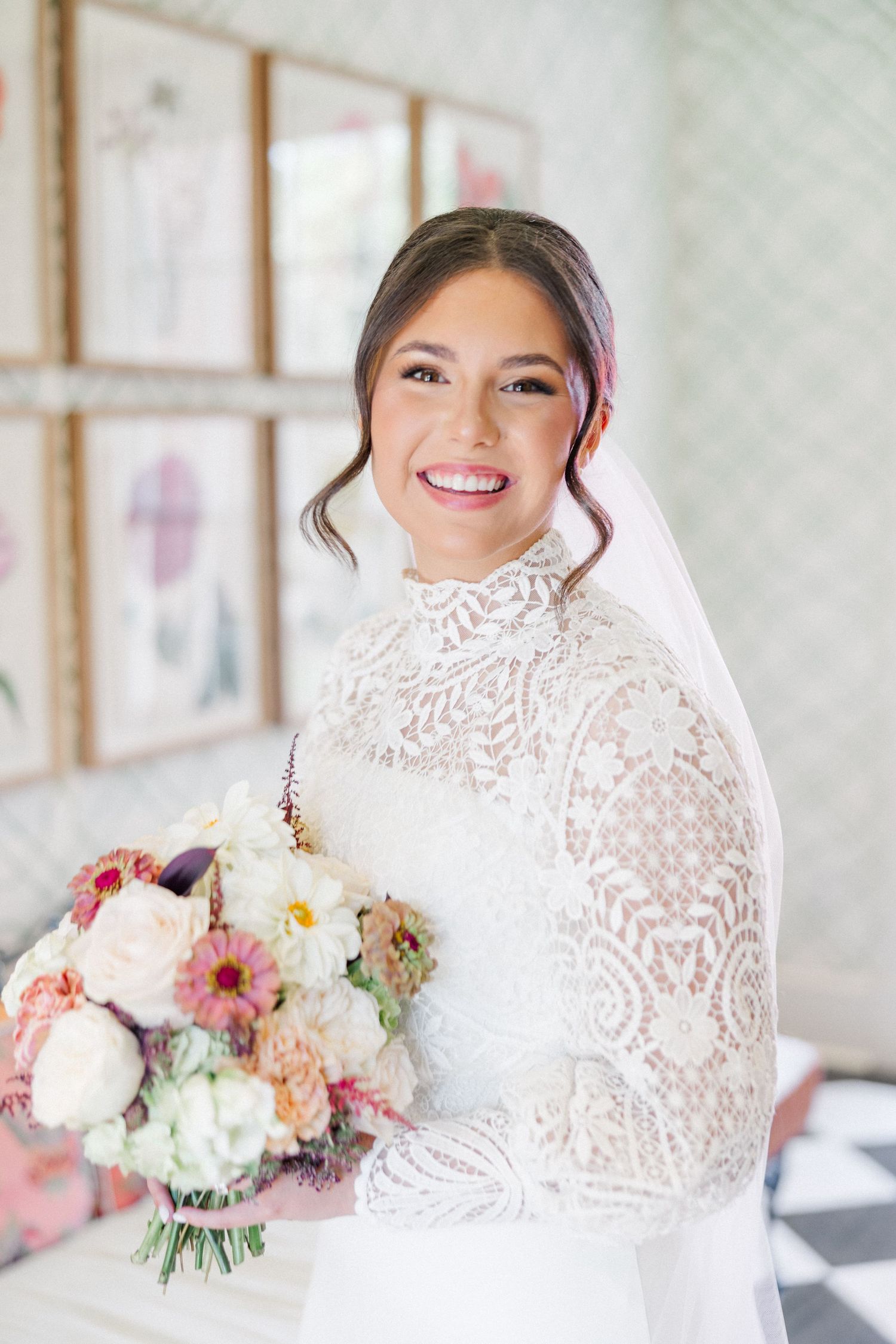 A bride wearing an elegant high-neck lace wedding dress smiles while holding a pink and white floral bouquet.