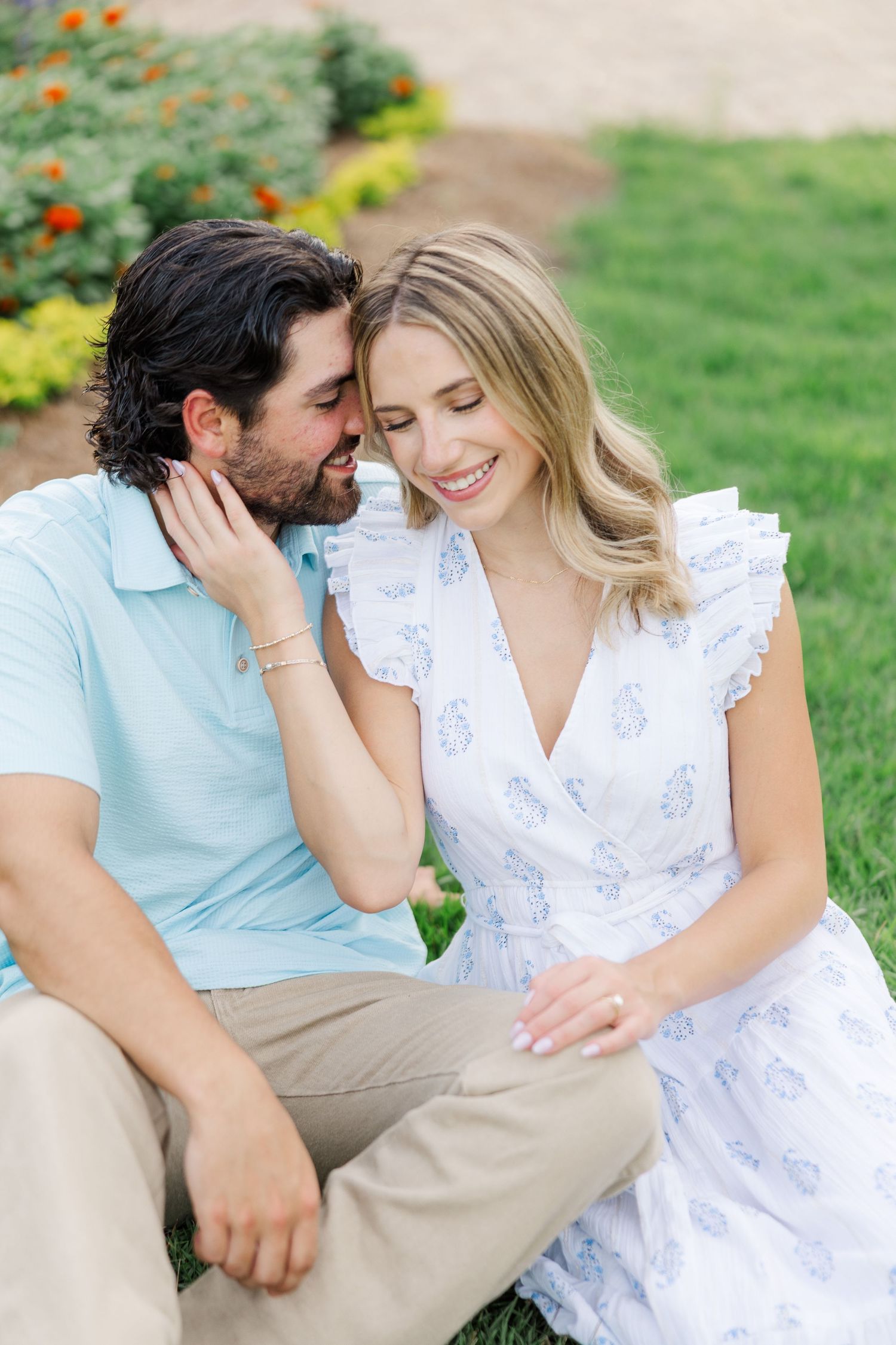 A couple in white and light blue outfits share tender moments outdoors on a sunny day in a garden setting.