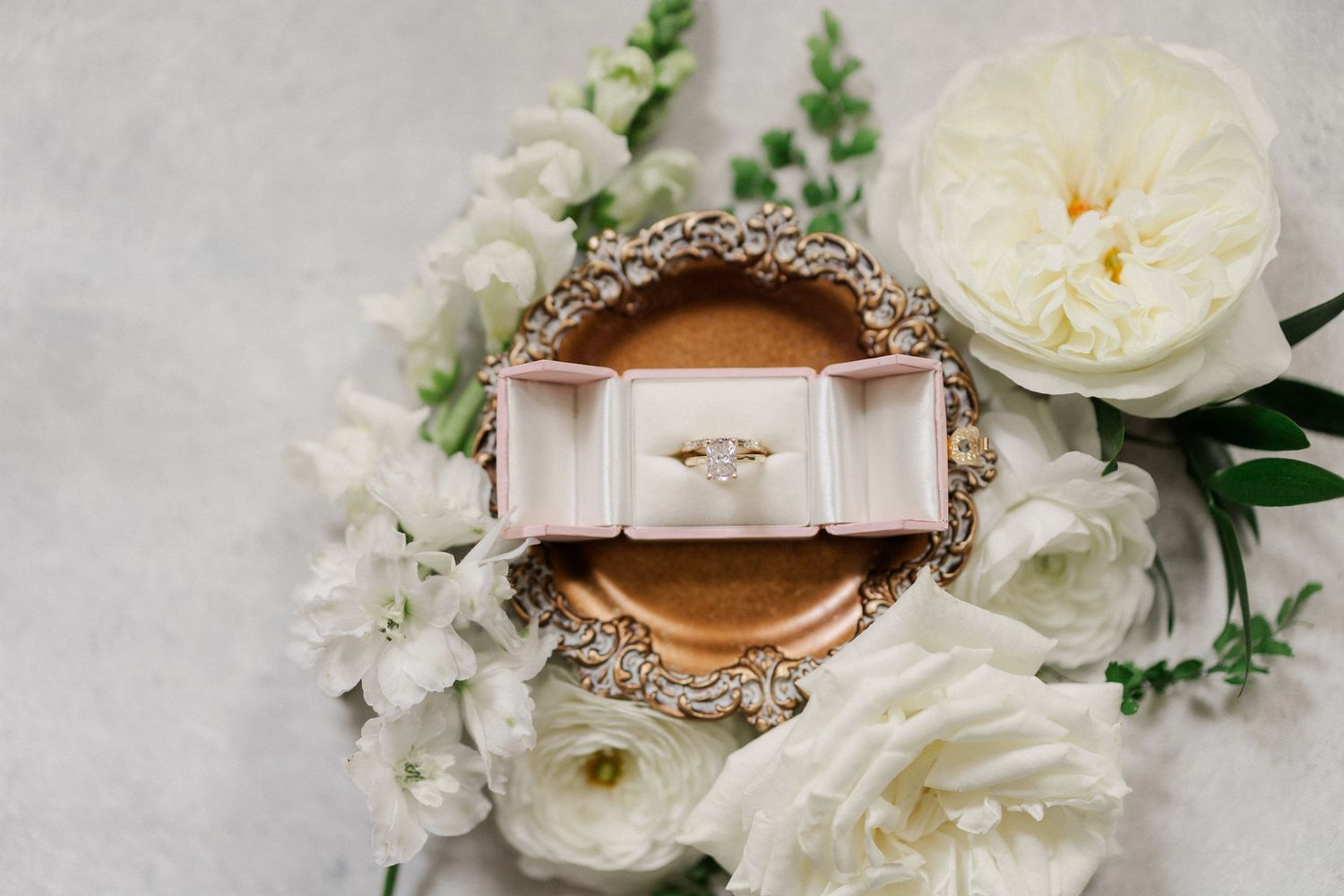 Gold engagement ring in velvet box surrounded by white peonies and ranunculus on gray background.
