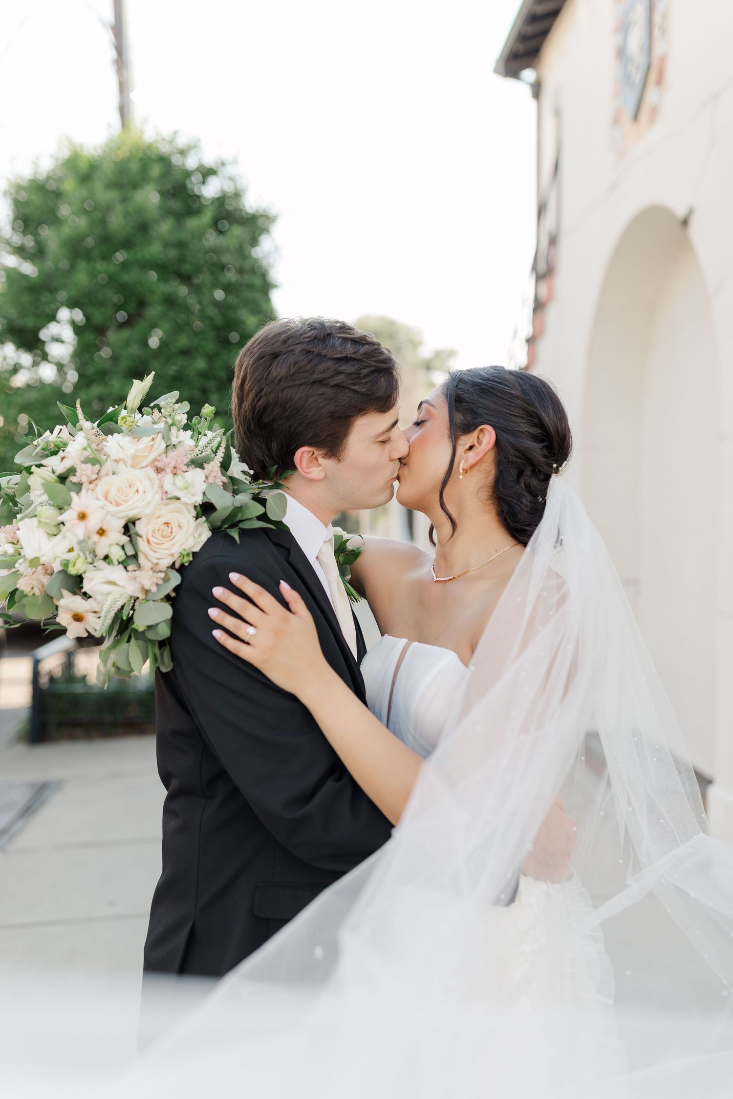 Tender wedding moment captured with flowing veil and white floral bouquet against light architecture.