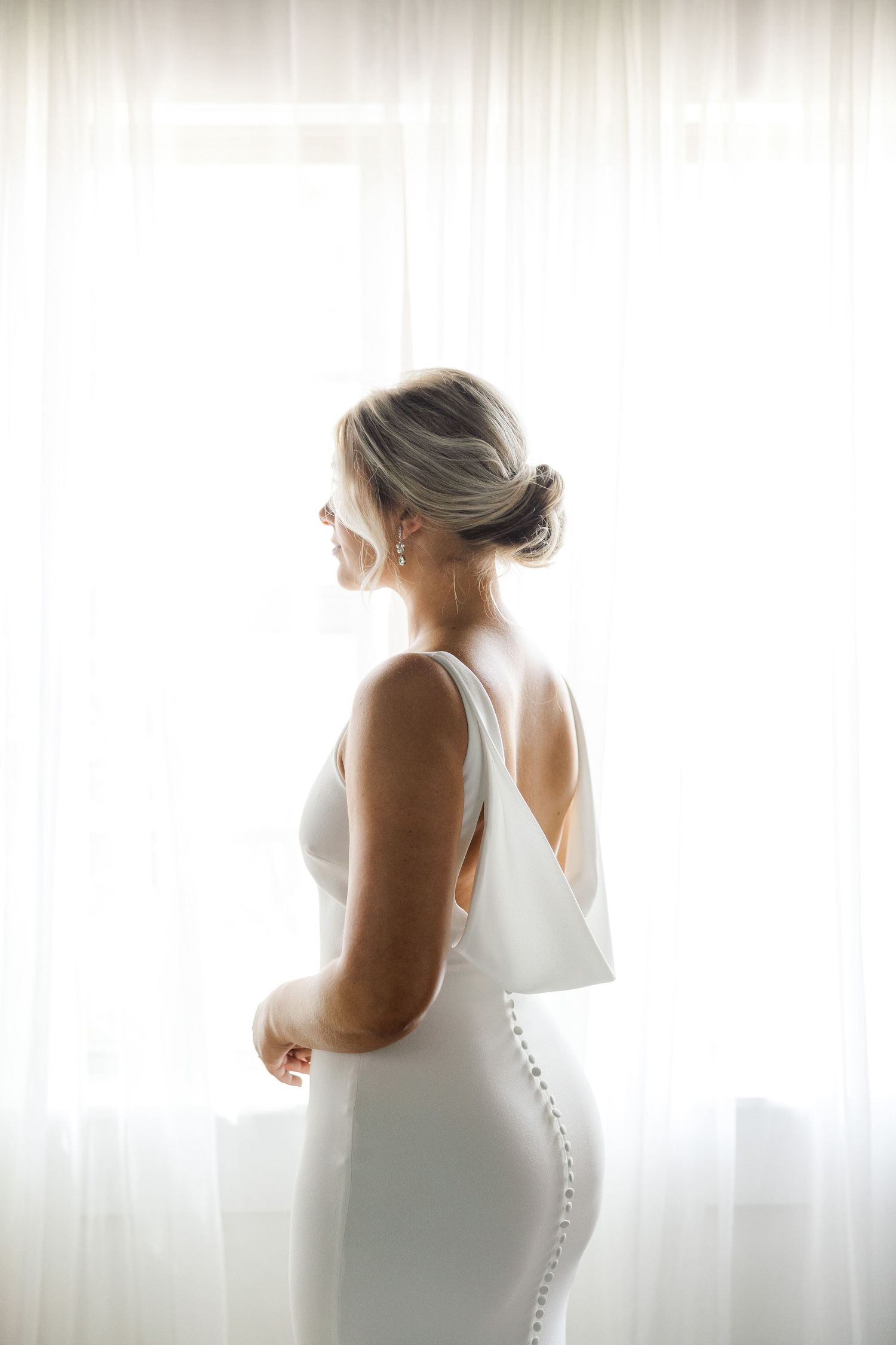 Bride in elegant fitted white gown poses by window in soft natural light.
