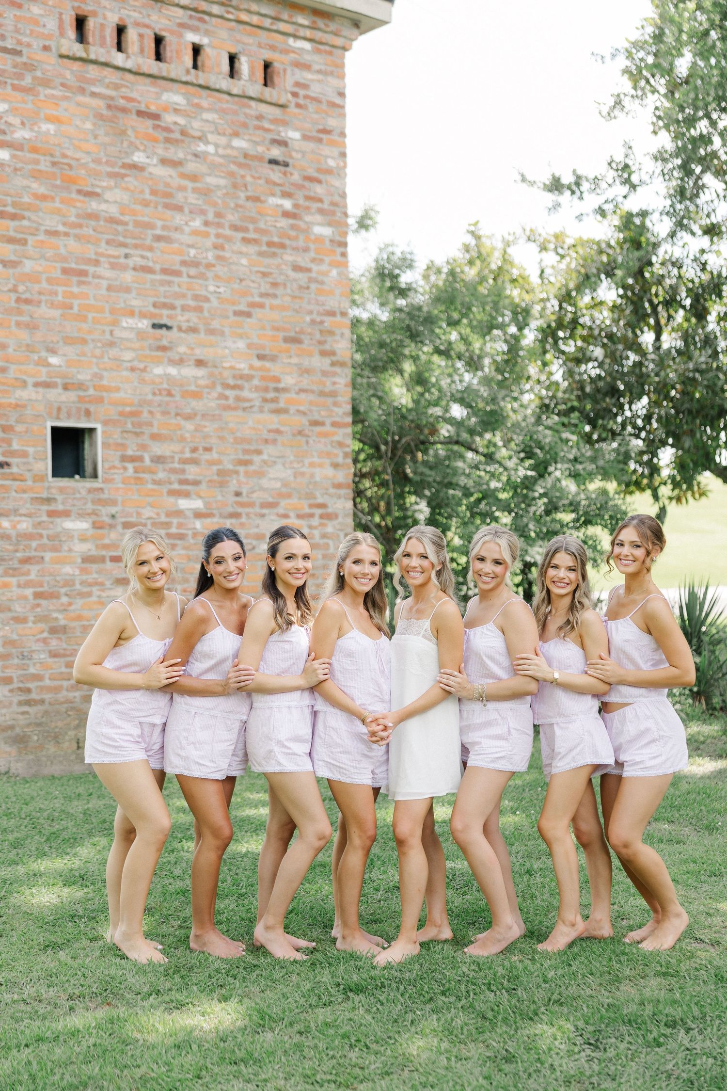 Eight bridesmaids in matching pink satin pajama sets posing barefoot on grass beside a rustic brick tower outdoors.