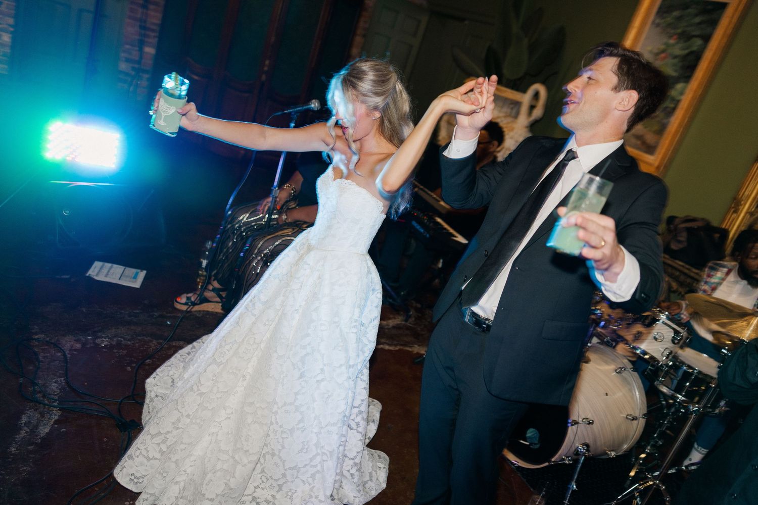 Bride and groom dance joyfully with champagne glasses raised near a live band at their wedding reception.