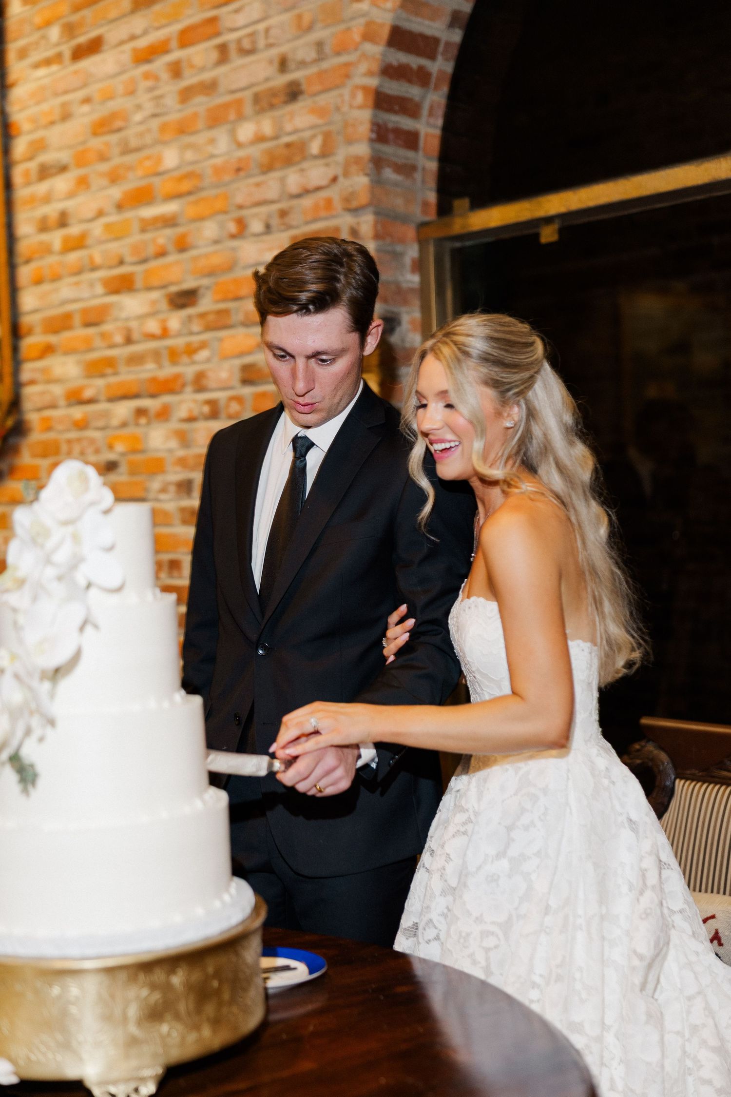 Bride and groom cut their white tiered wedding cake together, laughing in a brick-walled reception venue.