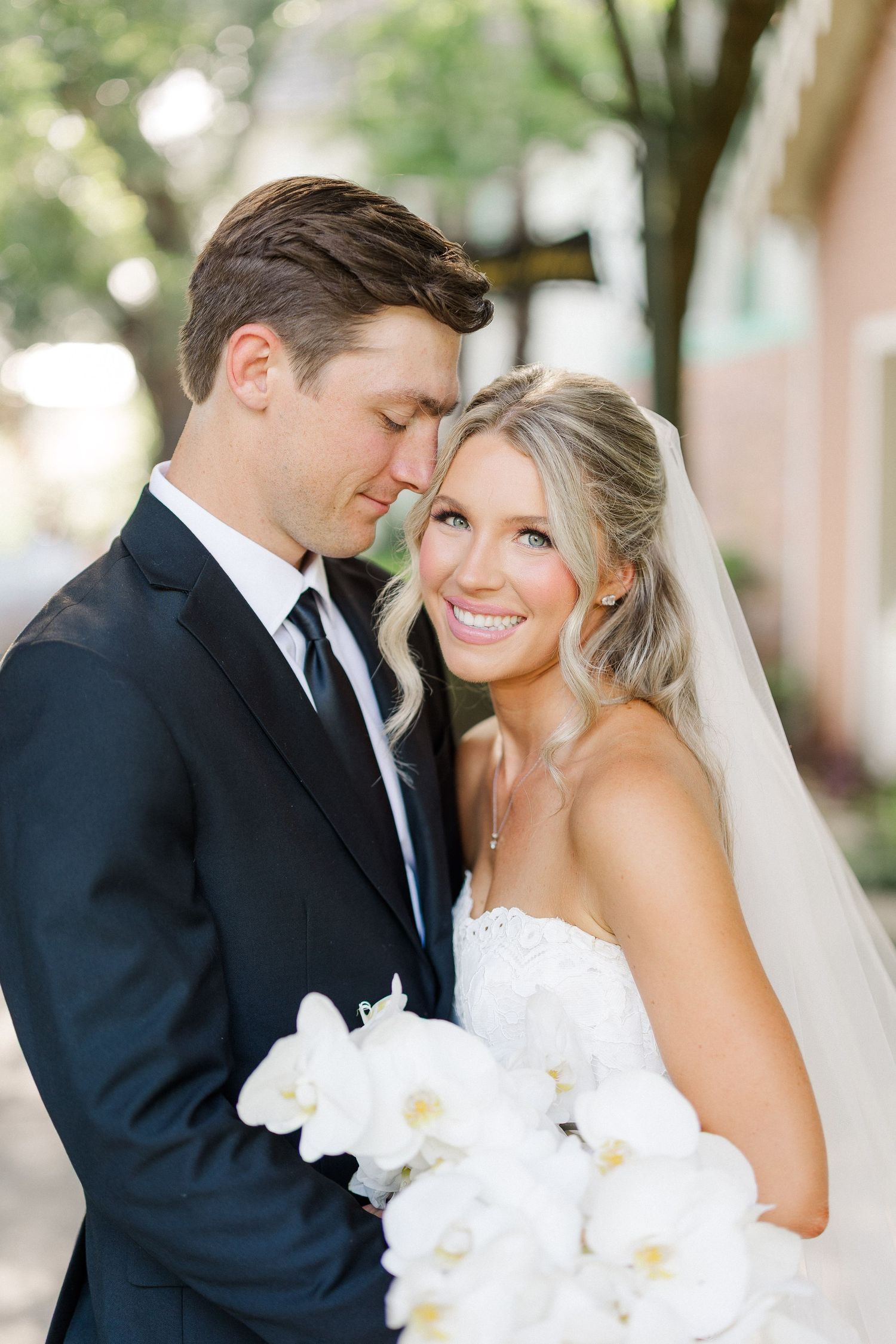 Bride and groom portrait, groom in navy suit holding white orchid bouquet, bride smiling in lace veil.