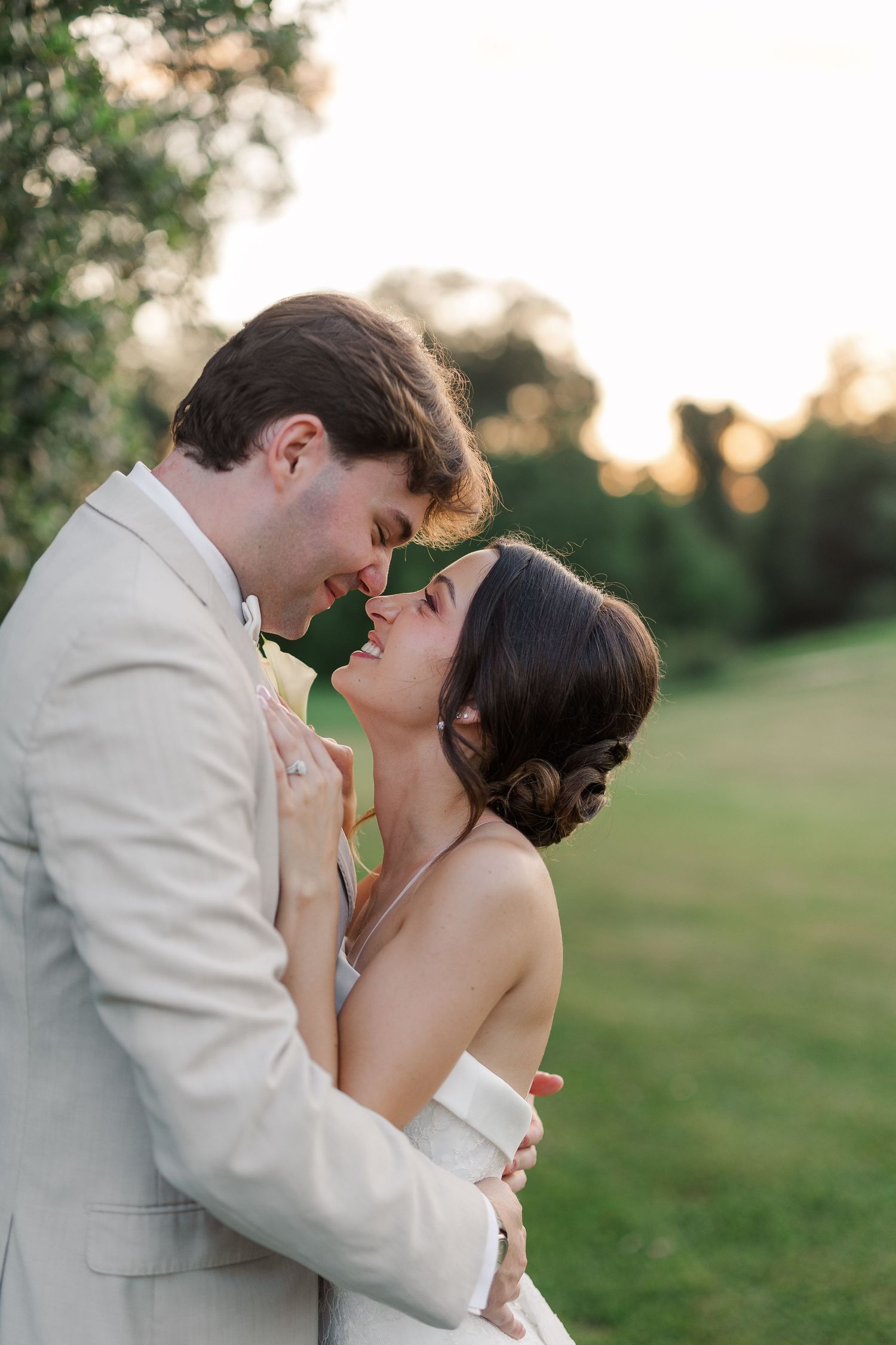 A bride and groom share a romantic almost-kiss embrace outdoors at golden hour, dressed in wedding attire.