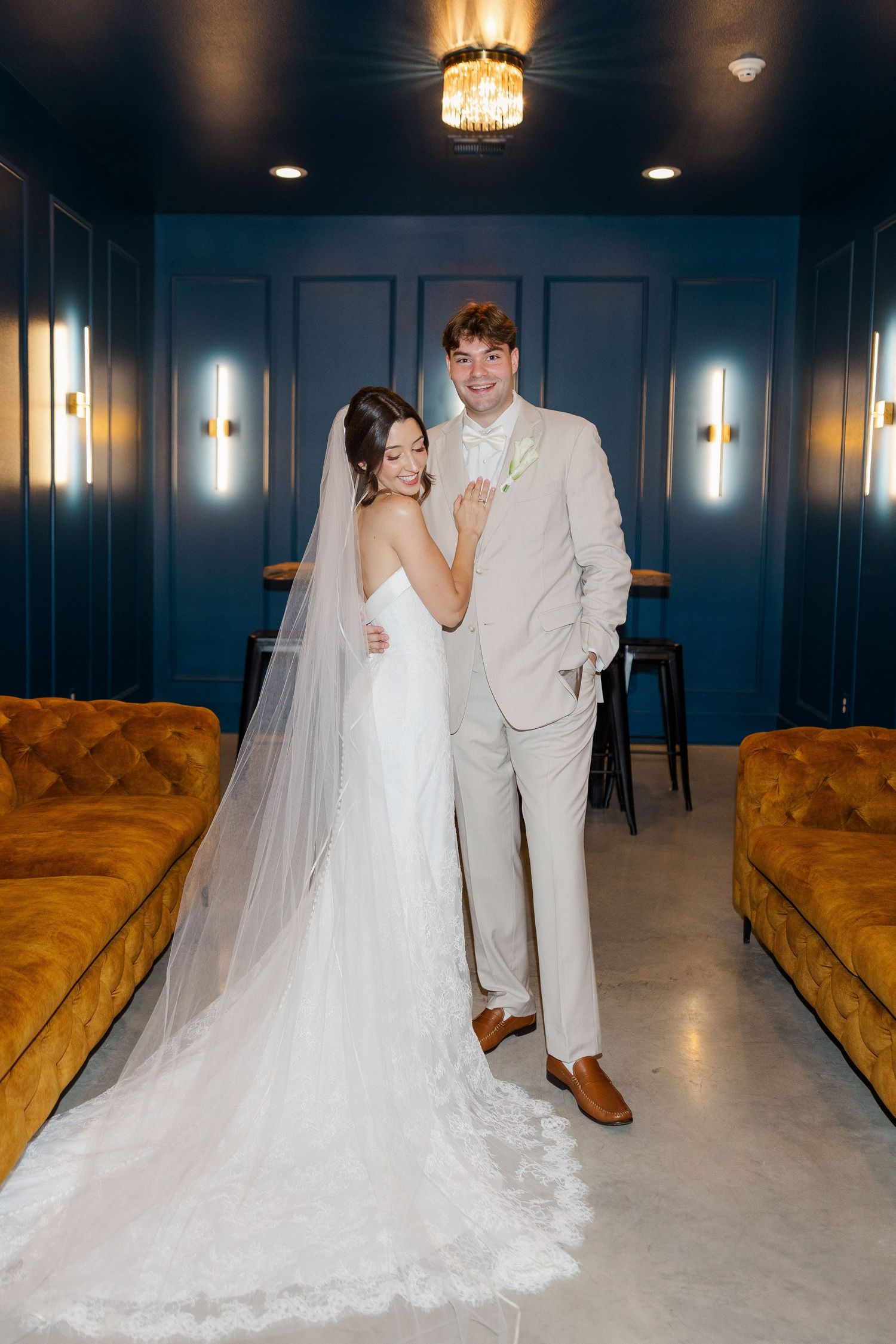 Bride in white gown with veil and groom in beige suit pose together in elegant blue room with gold accents.