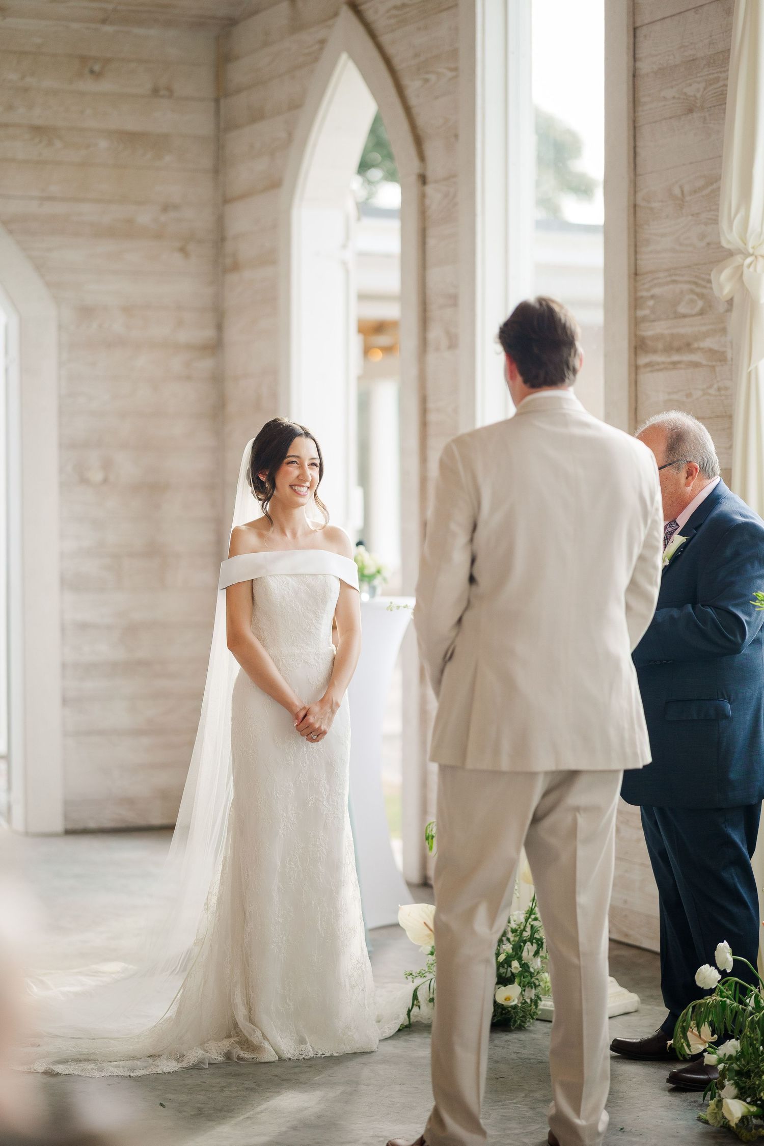 Radiant bride in off-shoulder wedding gown smiles at groom during intimate chapel wedding ceremony.