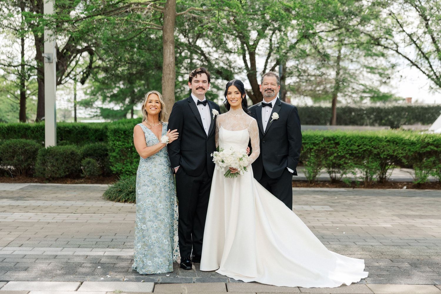 Wedding party of four poses outdoors, bride in white gown flanked by two men in tuxedos and woman in green gown.