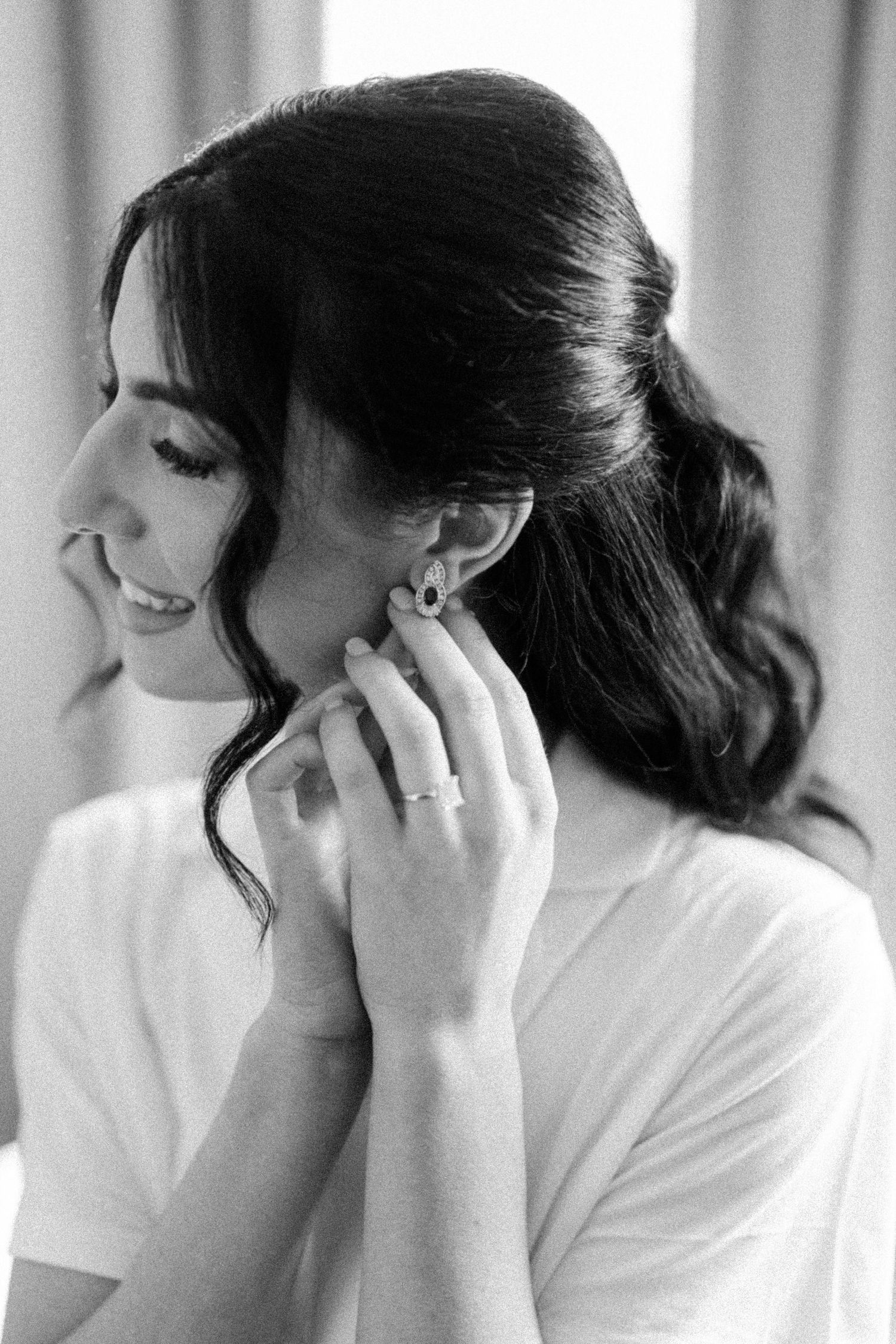 Black and white photo of a smiling bride putting on earrings, wearing a white dress and engagement ring.
