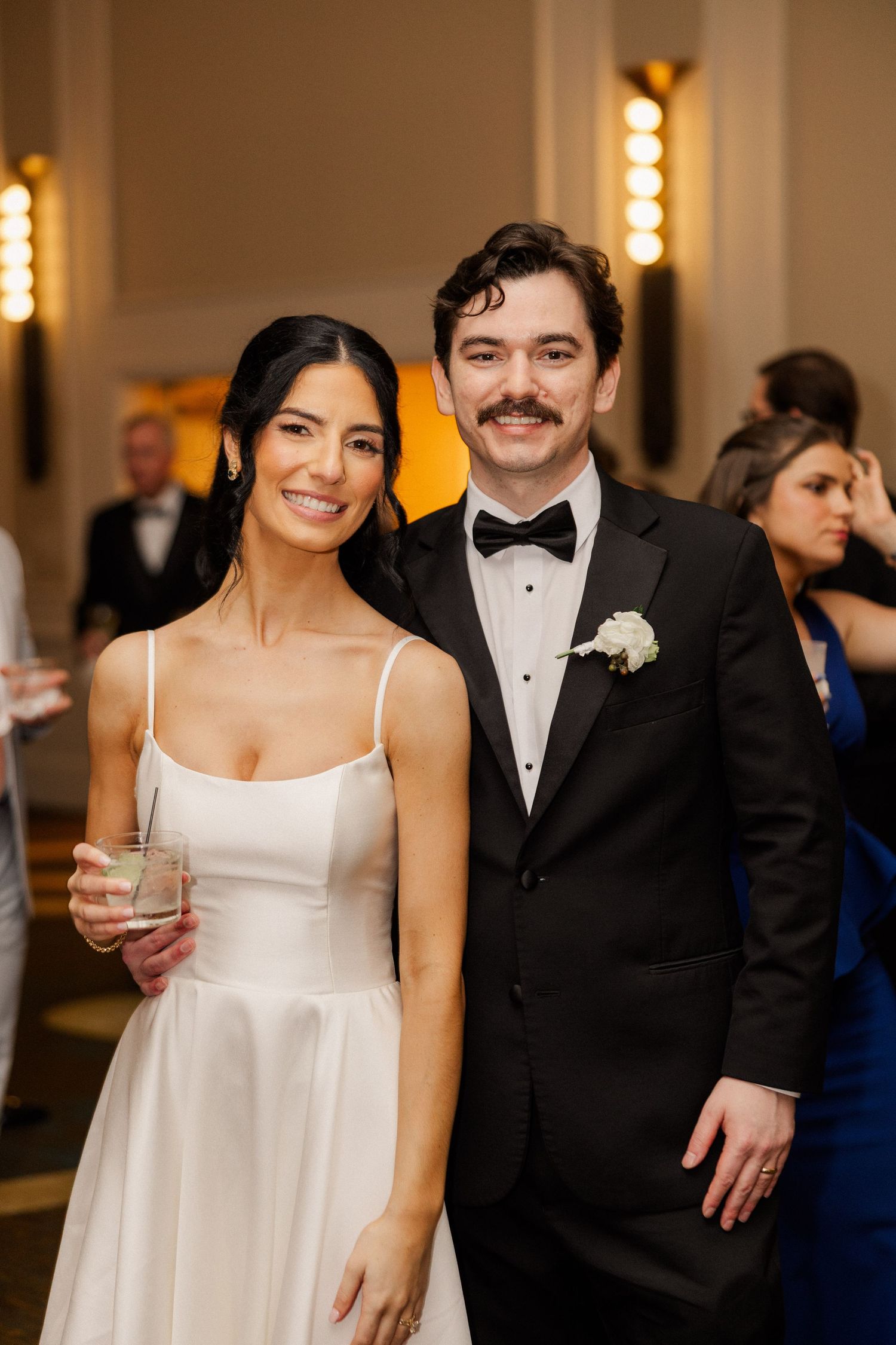 Bride in white gown and groom in black tuxedo with bow tie smile together at elegant wedding reception.