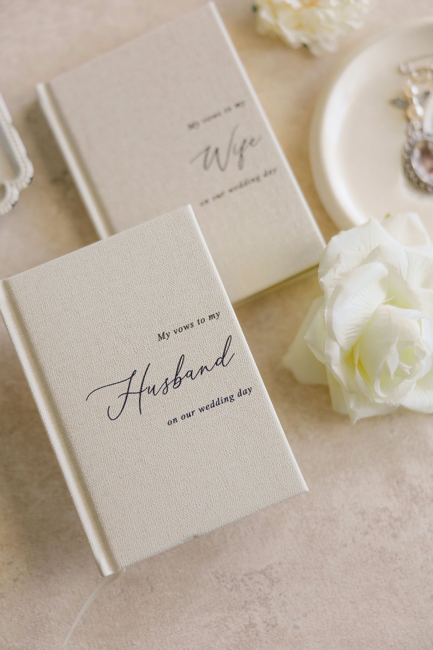 Two elegant wedding vow books in cream linen with calligraphy text and a white rose lying on a neutral surface.