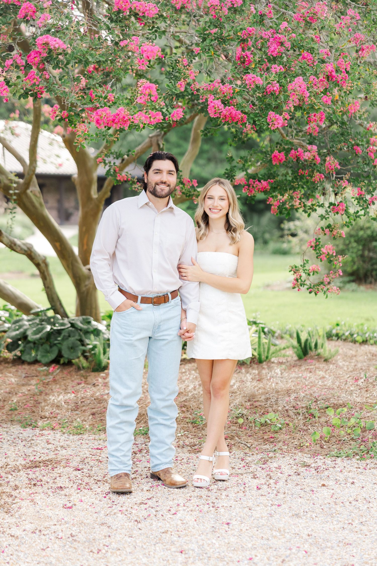 A couple poses together under a blooming pink crepe myrtle tree on a gravel path in a garden setting.