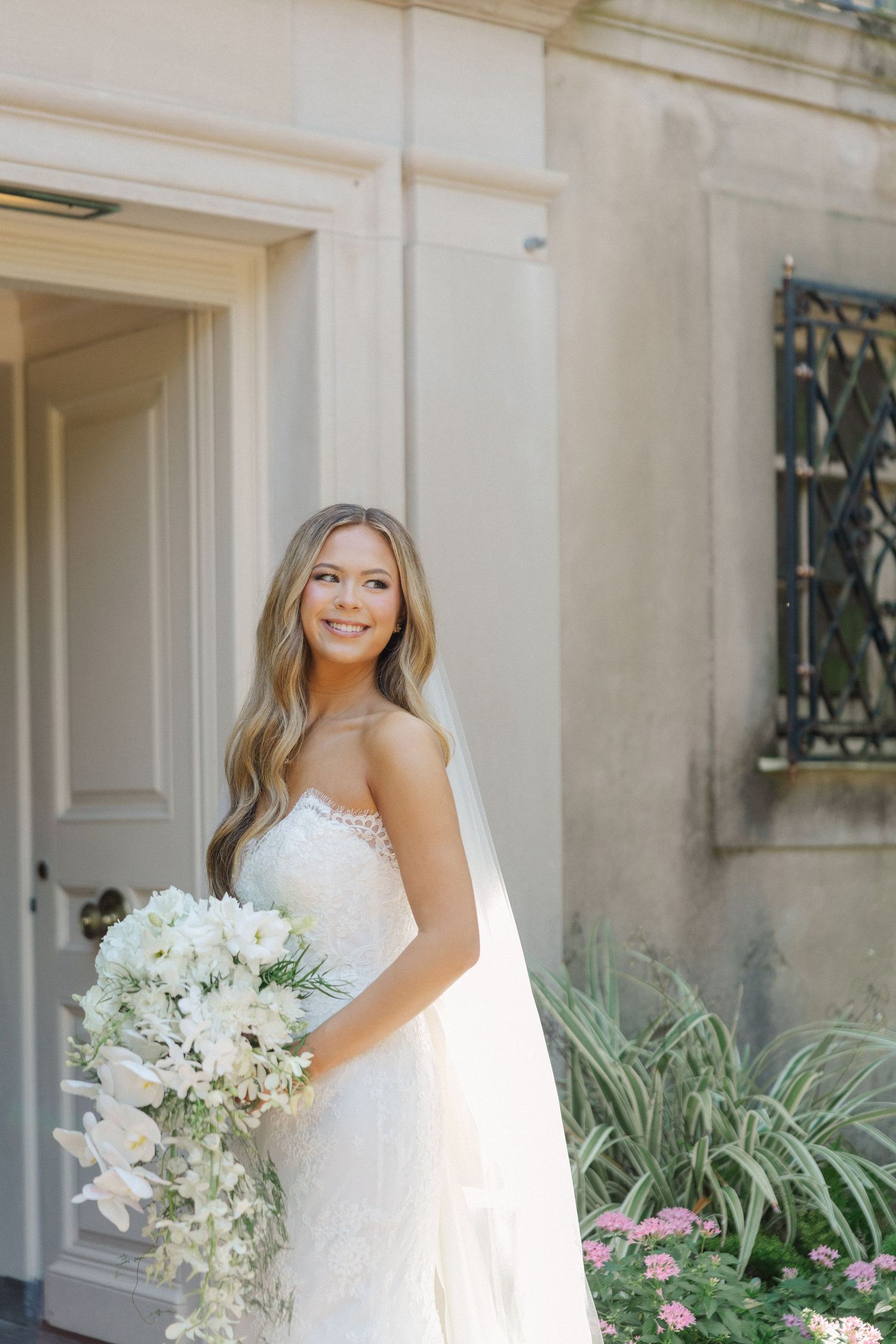 A bride in a white wedding dress holds a bouquet of white flowers while posing in front of a cream-colored building.