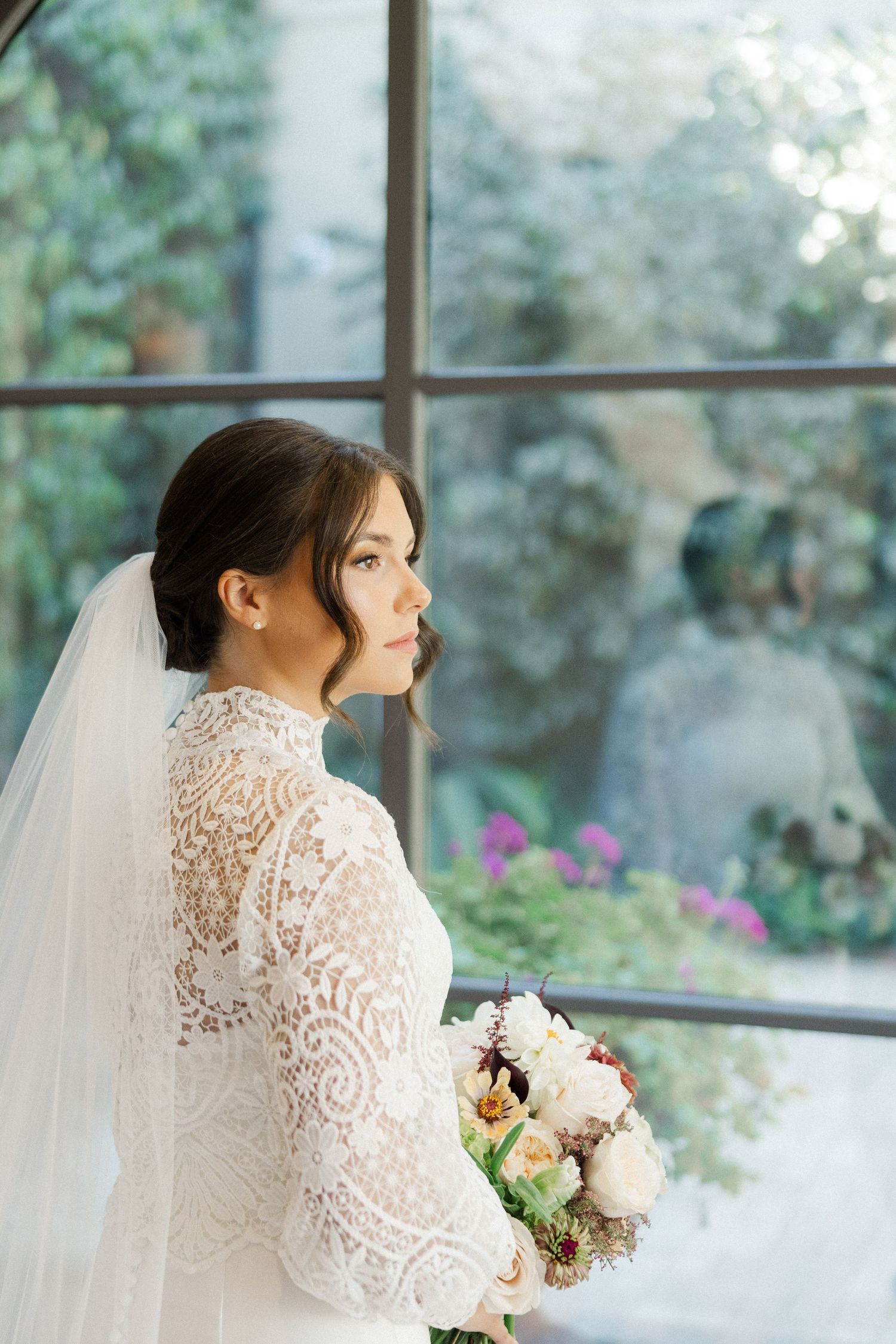 A bride in delicate lace holds a pastel bouquet while standing by a window with garden views.