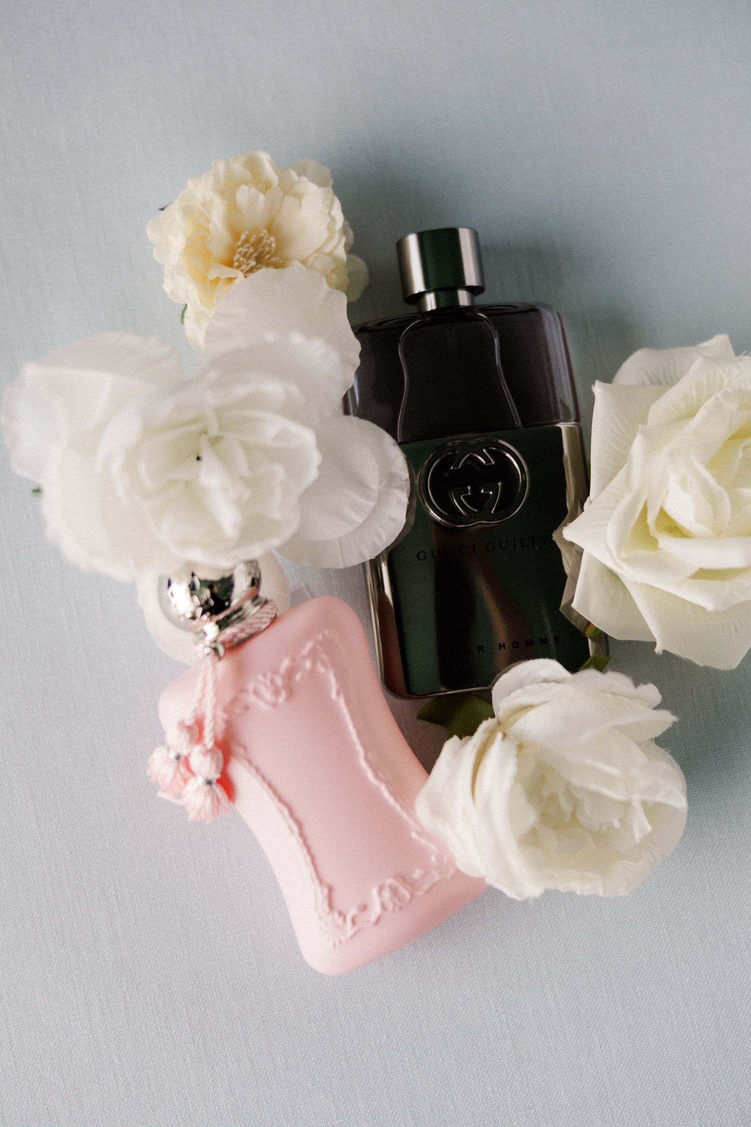 Elegant perfume bottles and white roses arranged with a pink decorative container on white surface.