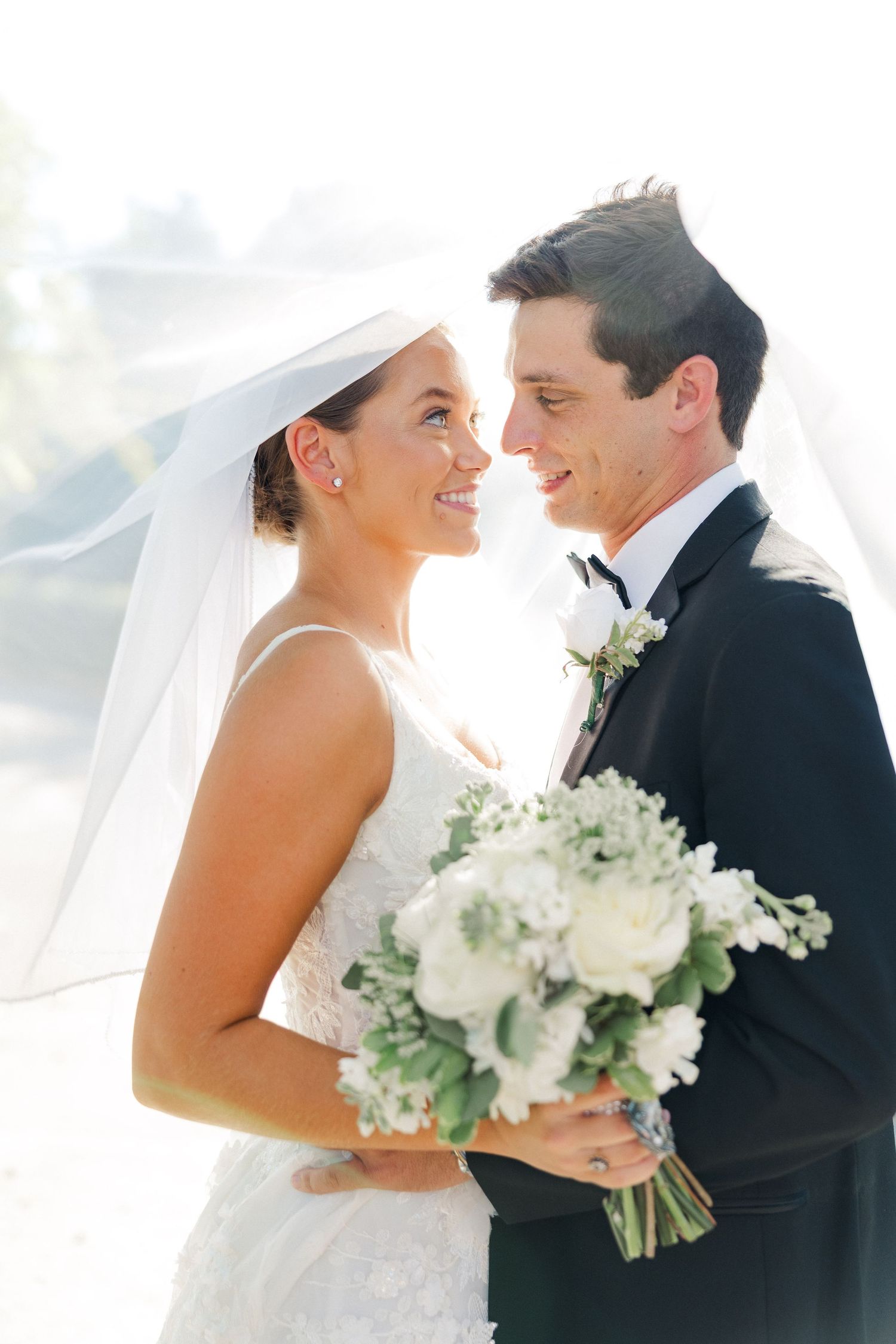 Intimate wedding couple moment with white floral bouquet and flowing veil in natural light.