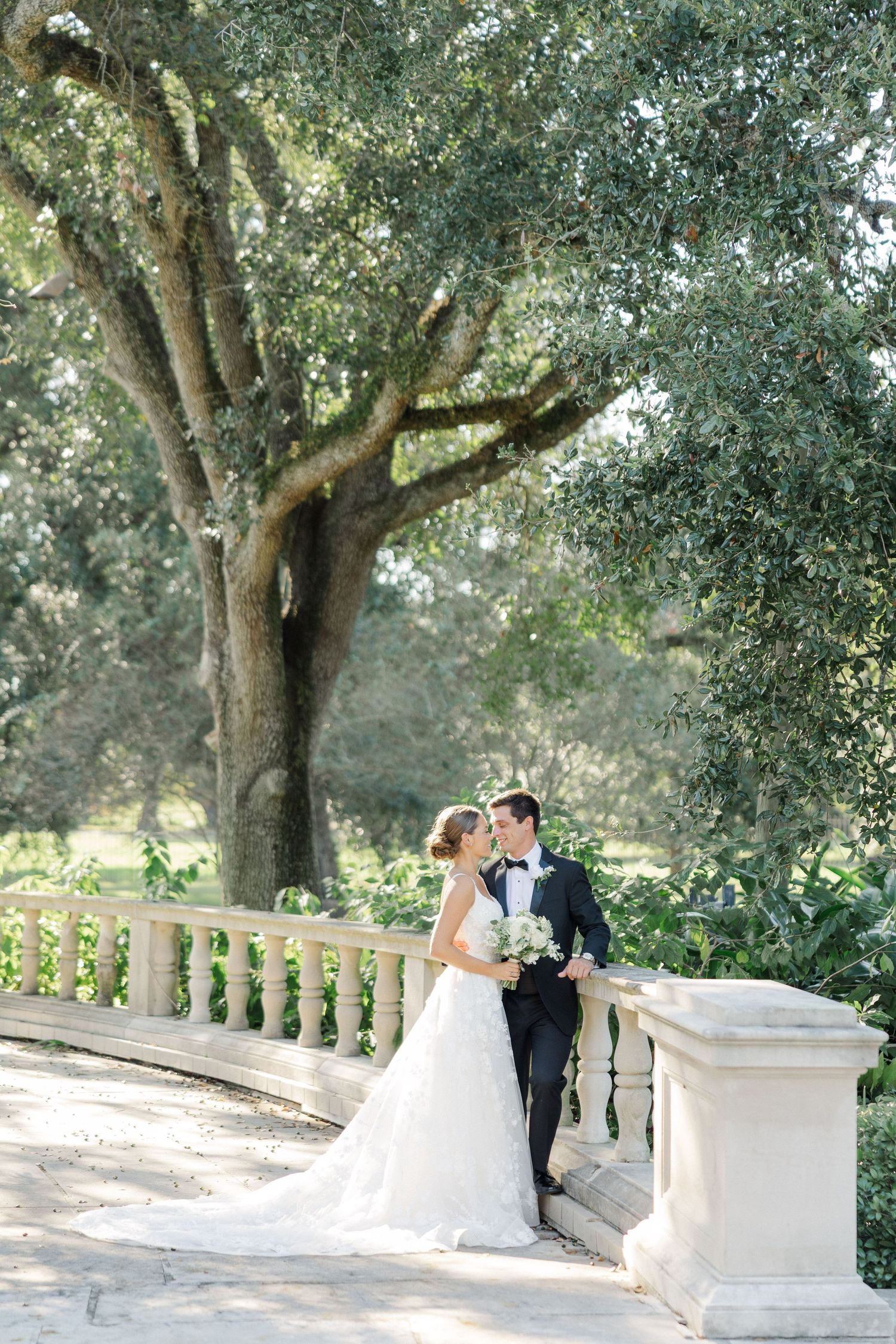 Newlyweds pose for romantic photos on a stone balustrade under oak trees.