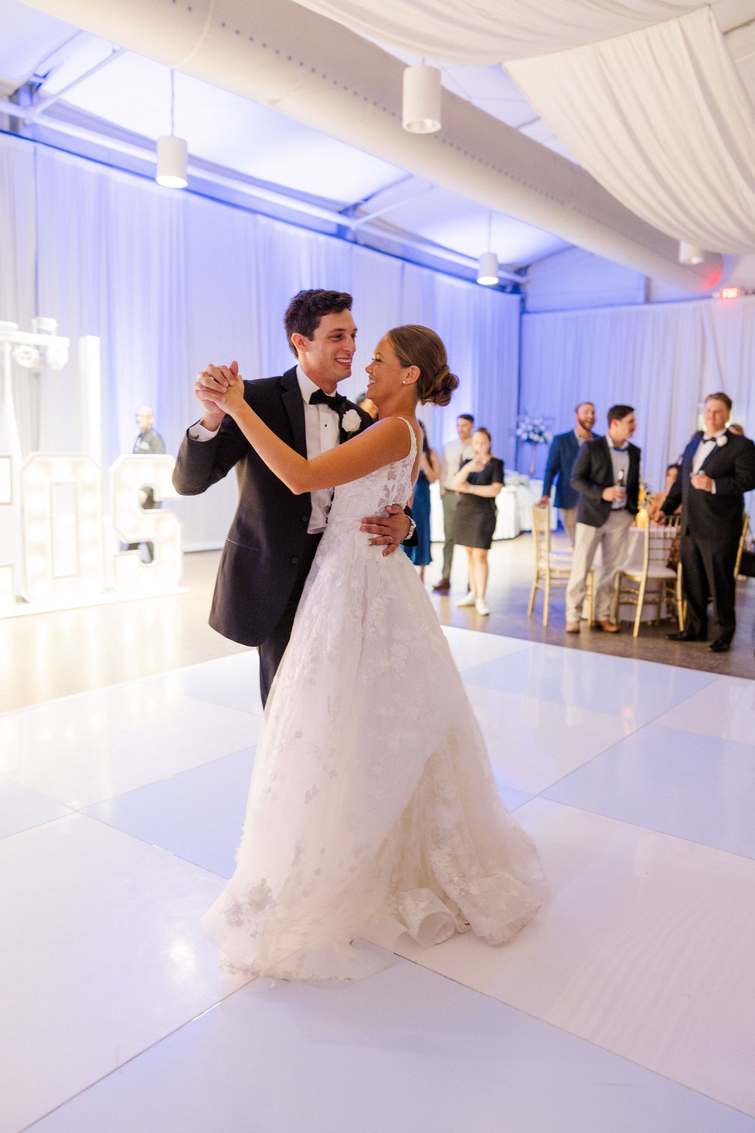 Couple shares their first dance in an elegant white ballroom with blue uplighting and draped fabric decor.
