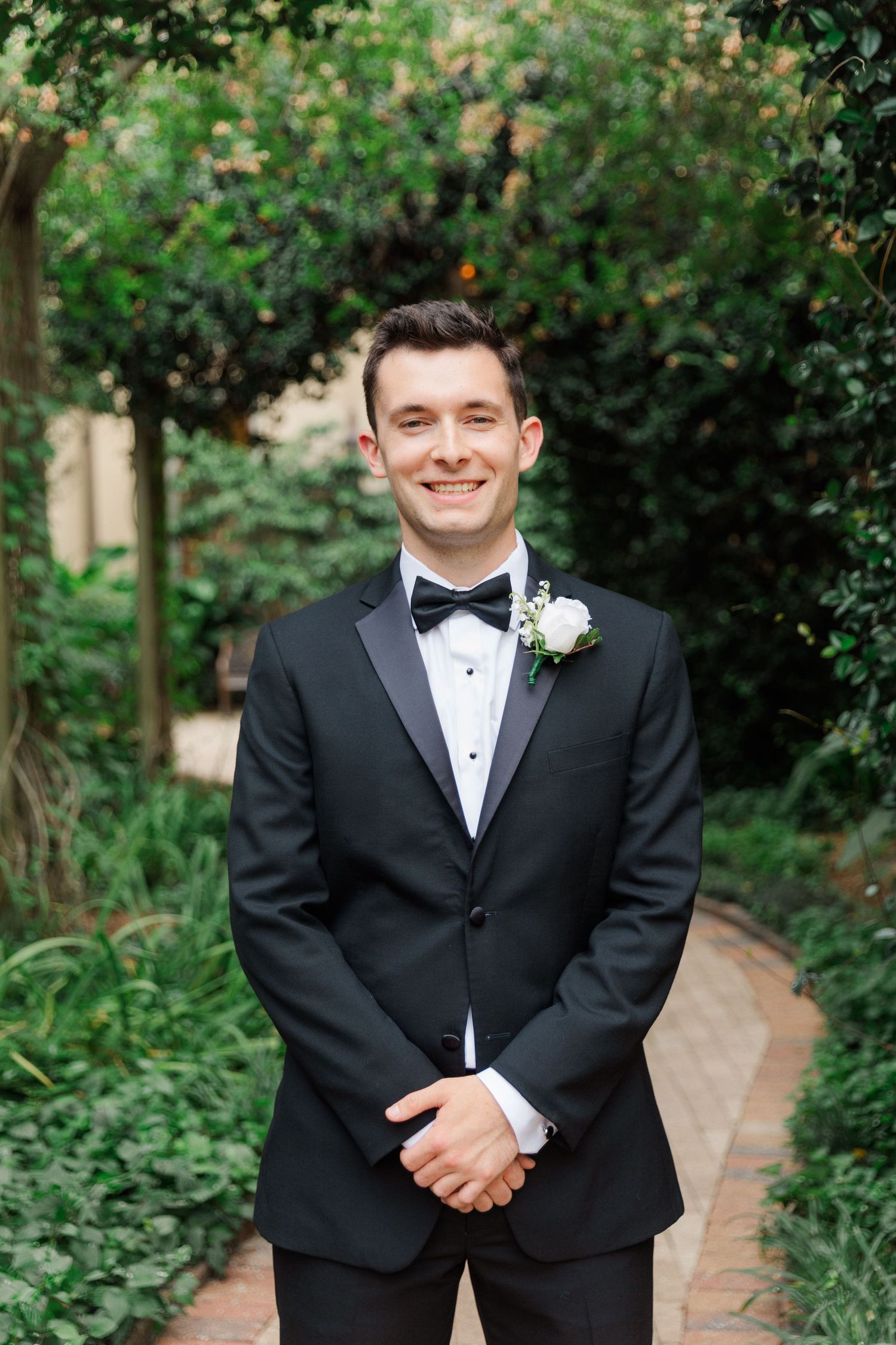Groom in classic black tuxedo and bow tie poses on garden path surrounded by lush greenery.
