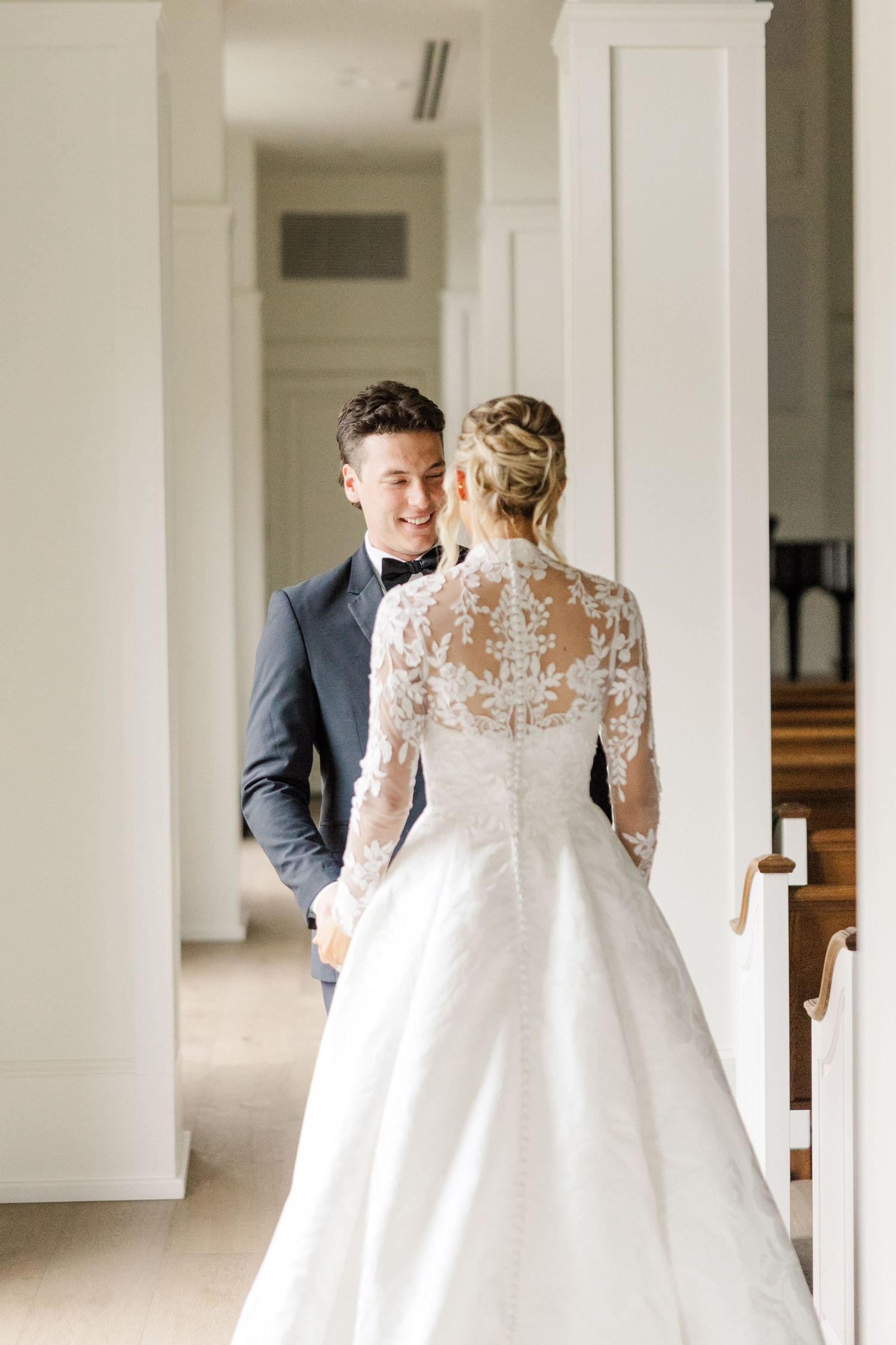 A bride in a lace-backed wedding gown stands with her groom in an elegant white hallway.