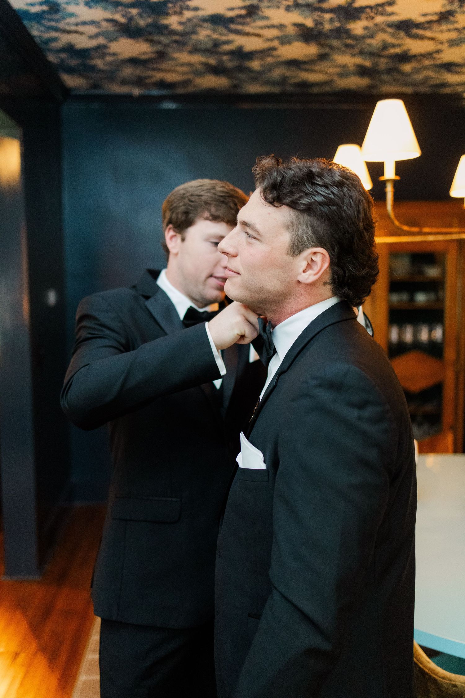 Two men in black tuxedos share an emotional moment in a dimly lit room with warm lighting.