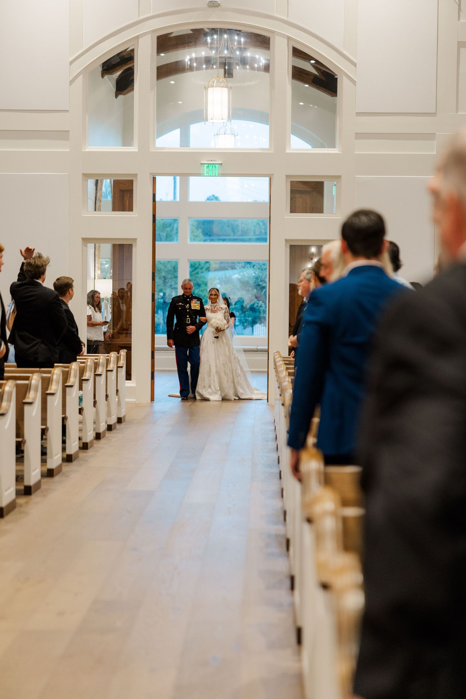 Wedding ceremony aisle in an elegant white chapel with wooden pews and guests seated.