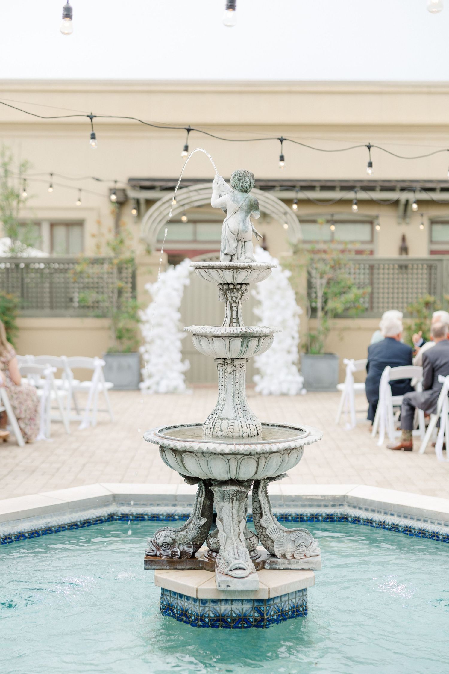 Ornate tiered fountain in courtyard with white floral arch in background.