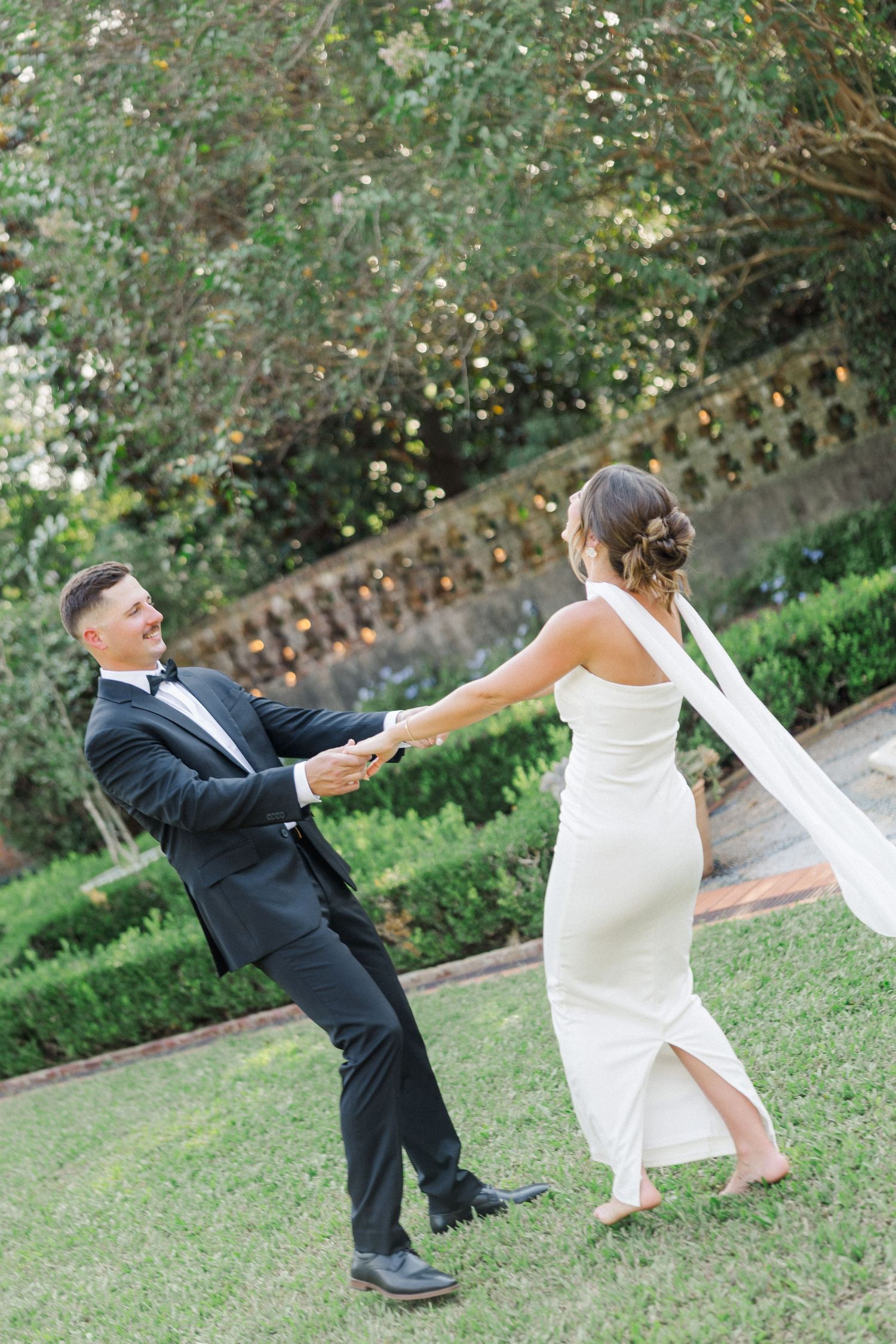 A couple in wedding attire playfully dances together in a garden setting.